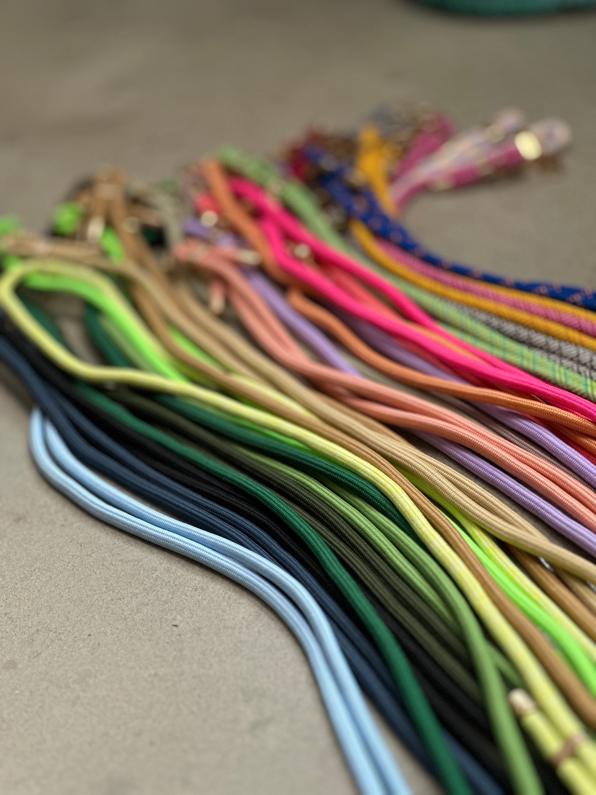 Eine Nahaufnahme von mehreren LEY! Handykette - Roséfarbene Kordeln und Schlüsselbänder, die in Reihen angeordnet sind und deren leuchtende Farben - wie Schwarz, Blau, Grün, Pink, Orange, Hellbraun und mehr - hervorheben.