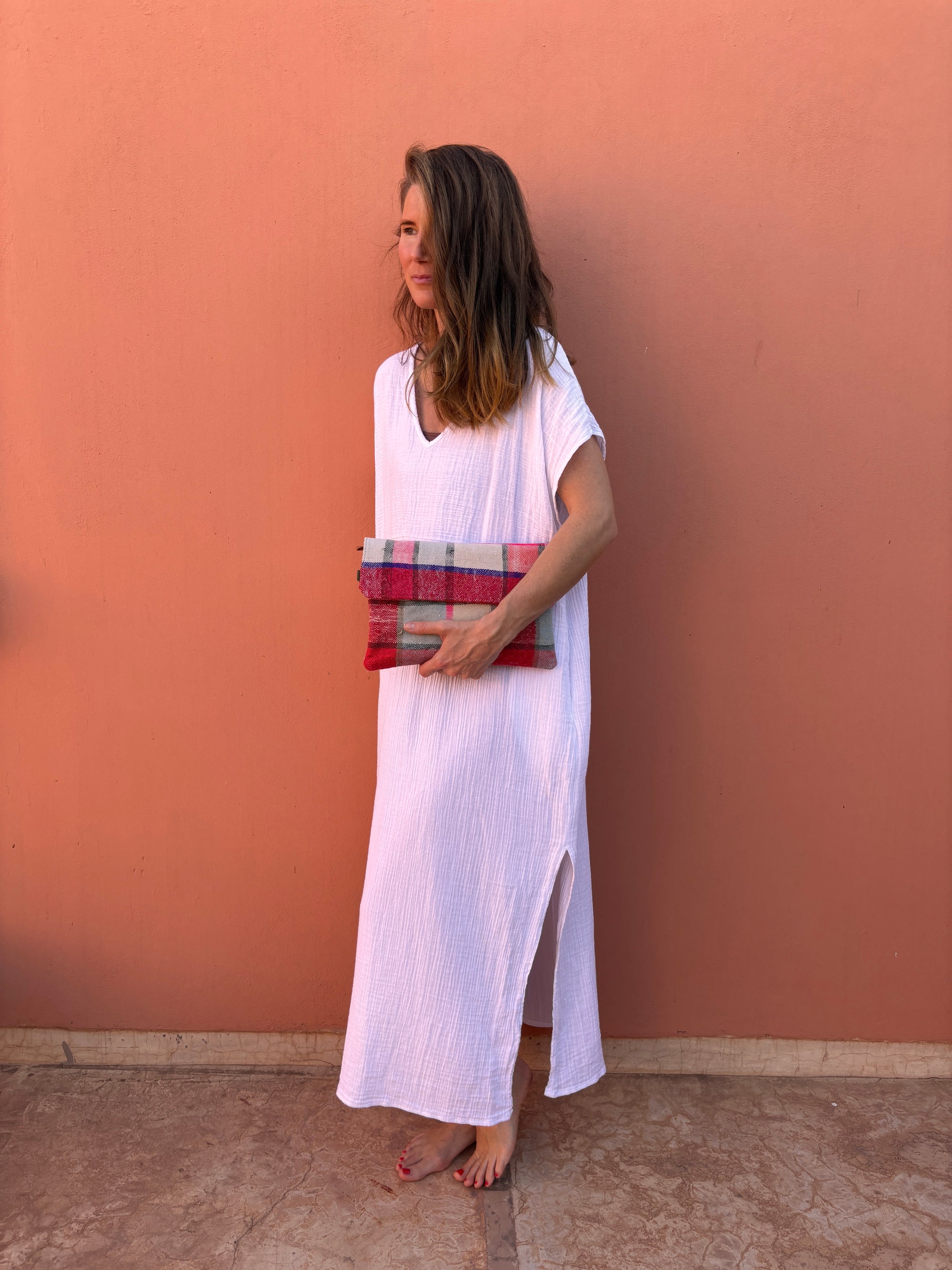 Eine Frau in einem weiten weißen Kleid steht barfuß vor einer pfirsichfarbenen Wand, hält die LEY! Vintage Clutch in der Hand und schaut mit einem leichten Lächeln zur Seite.