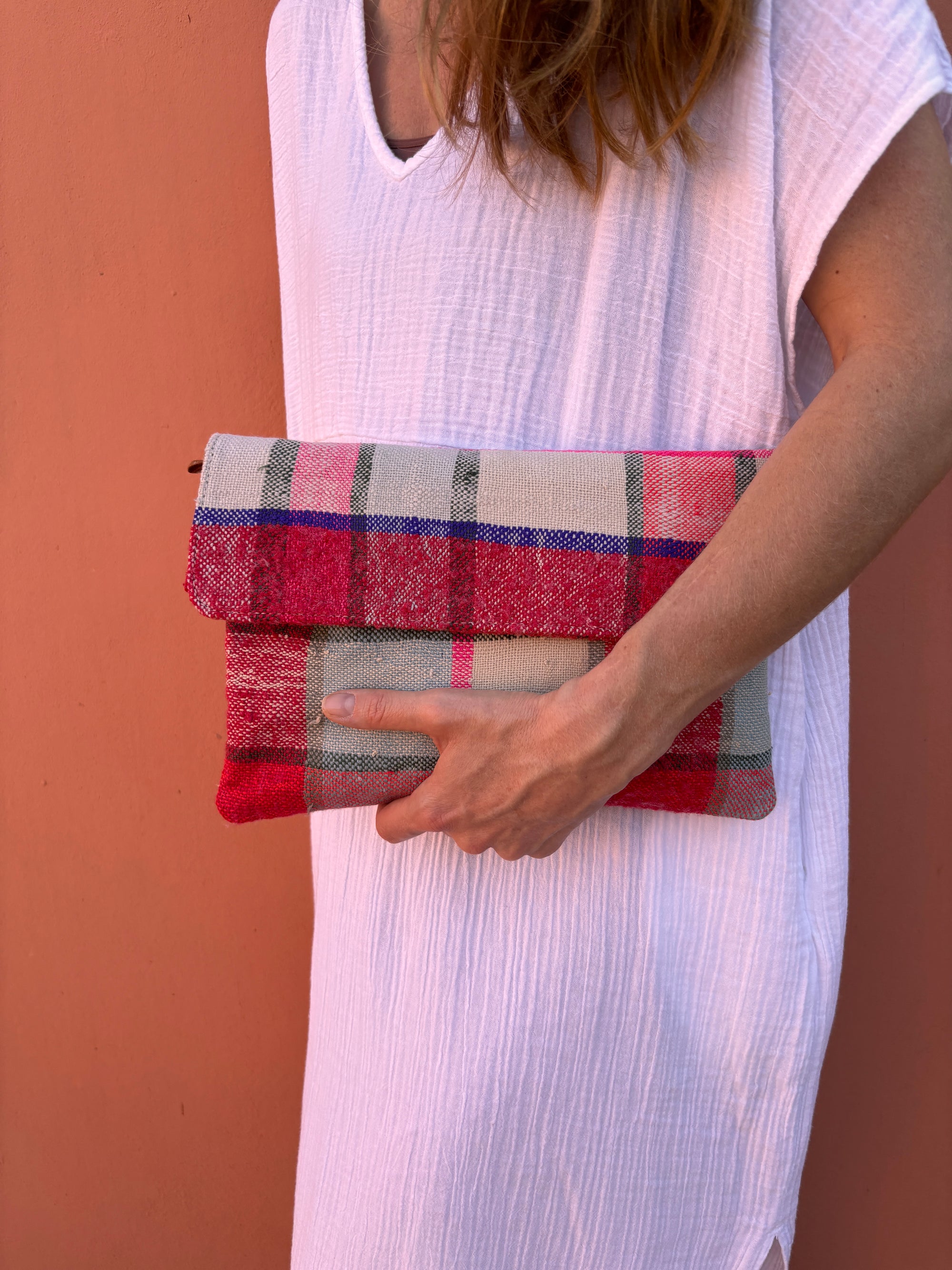Eine Person in einem weißen, strukturierten Kleid hält eine LEY! Vintage Clutch mit einem rot, rosa und grau karierten Muster aus Wolle vor einer pfirsichfarbenen Wand.