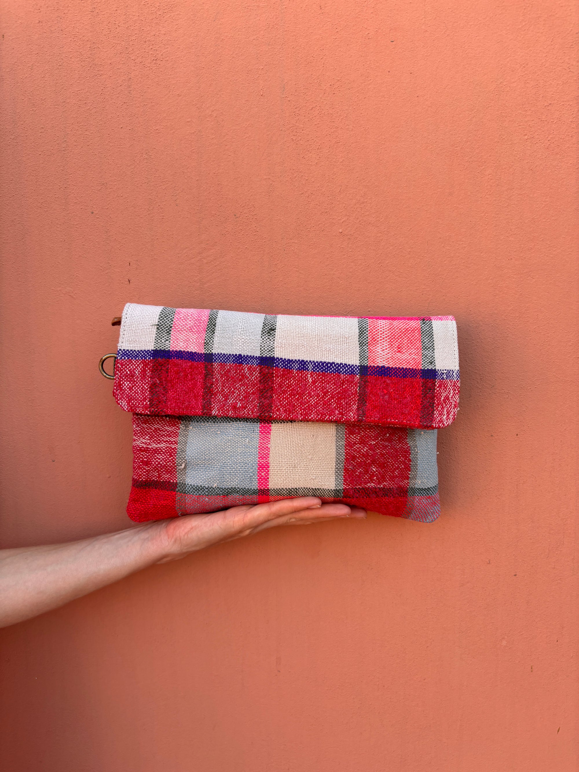 Eine Hand hält die LEY! Vintage Clutch, handgefertigt aus Wollstoffen mit einem auffälligen Karomuster in Rot, Rosa, Weiß, Blau und Grau, mit einer Klappe zum Umklappen und vor einer einfarbigen pfirsichfarbenen Wand.