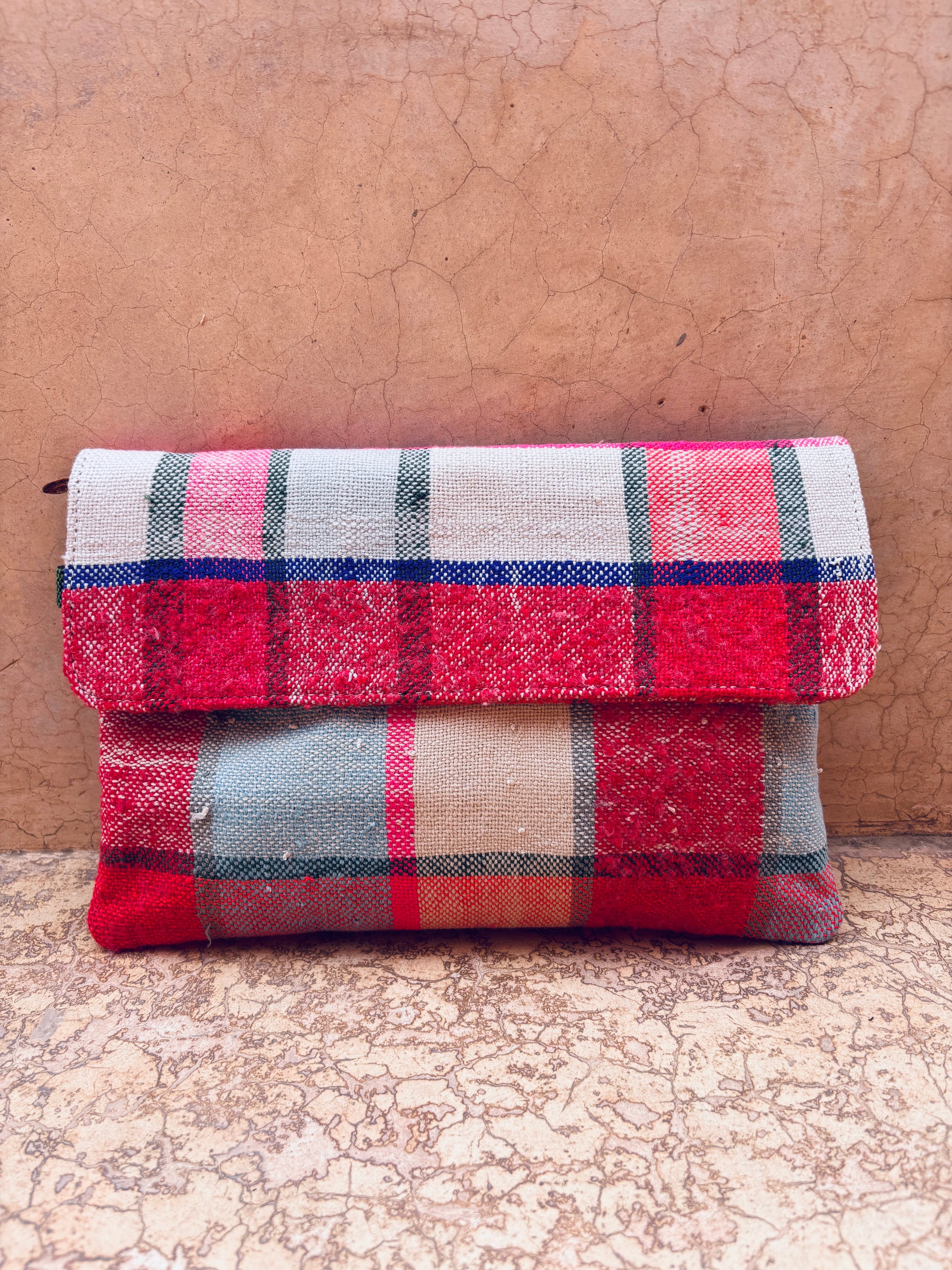 Die LEY! Vintage Clutch mit rot-weiß-grün-blauem Karomuster aus Upcycling Wolle steht vor einer rissigen, strukturierten beigen Wand auf dem Boden.