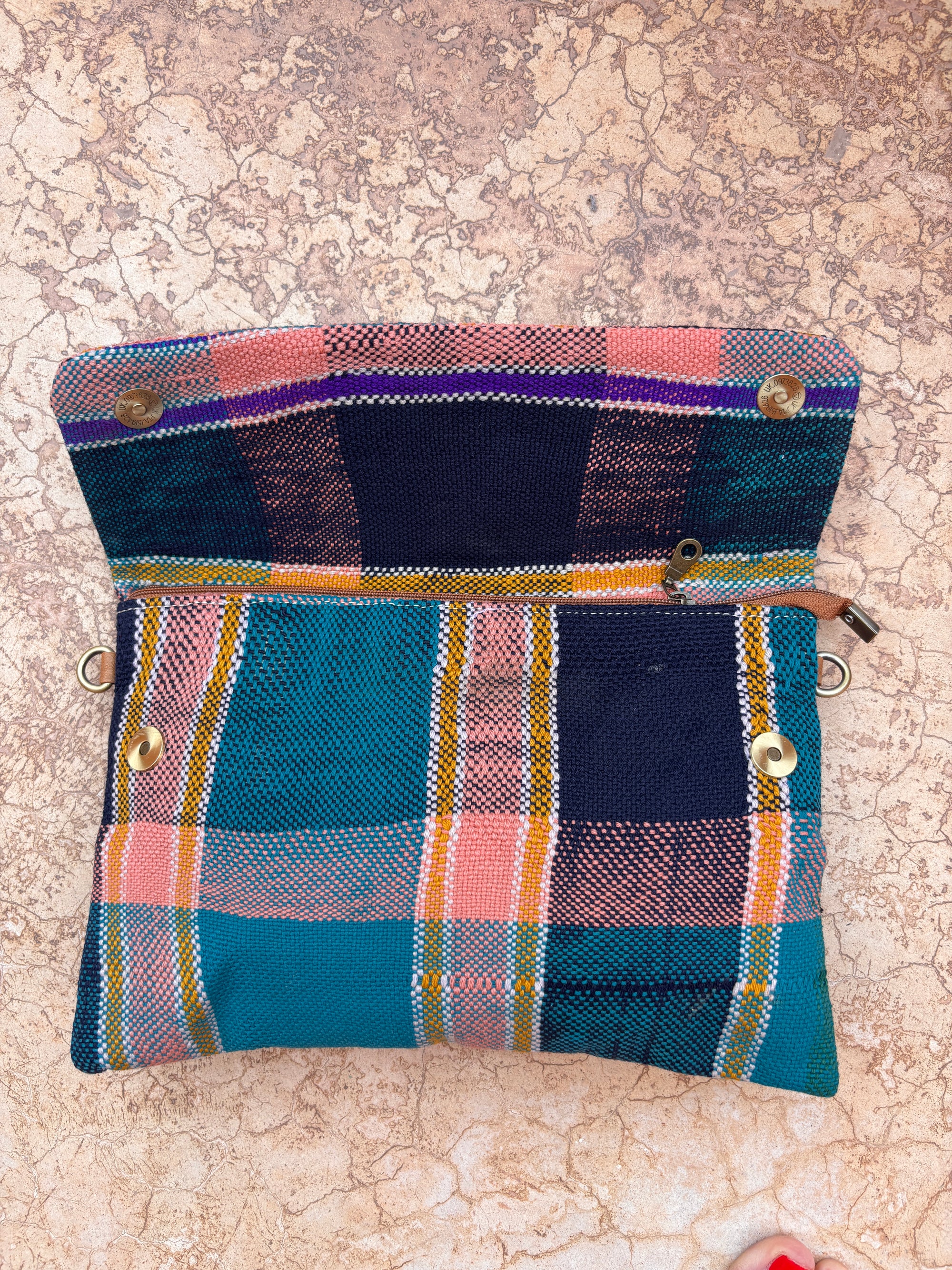 Die LEY! Vintage Clutch aus bunt kariertem Wollstoff mit blaugrünen, marineblauen, gelben und rosafarbenen Streifen und goldenen Druckknöpfen liegt offen auf einer Steinoberfläche. Aus der Ecke lugt ein rosa Gegenstand hervor.