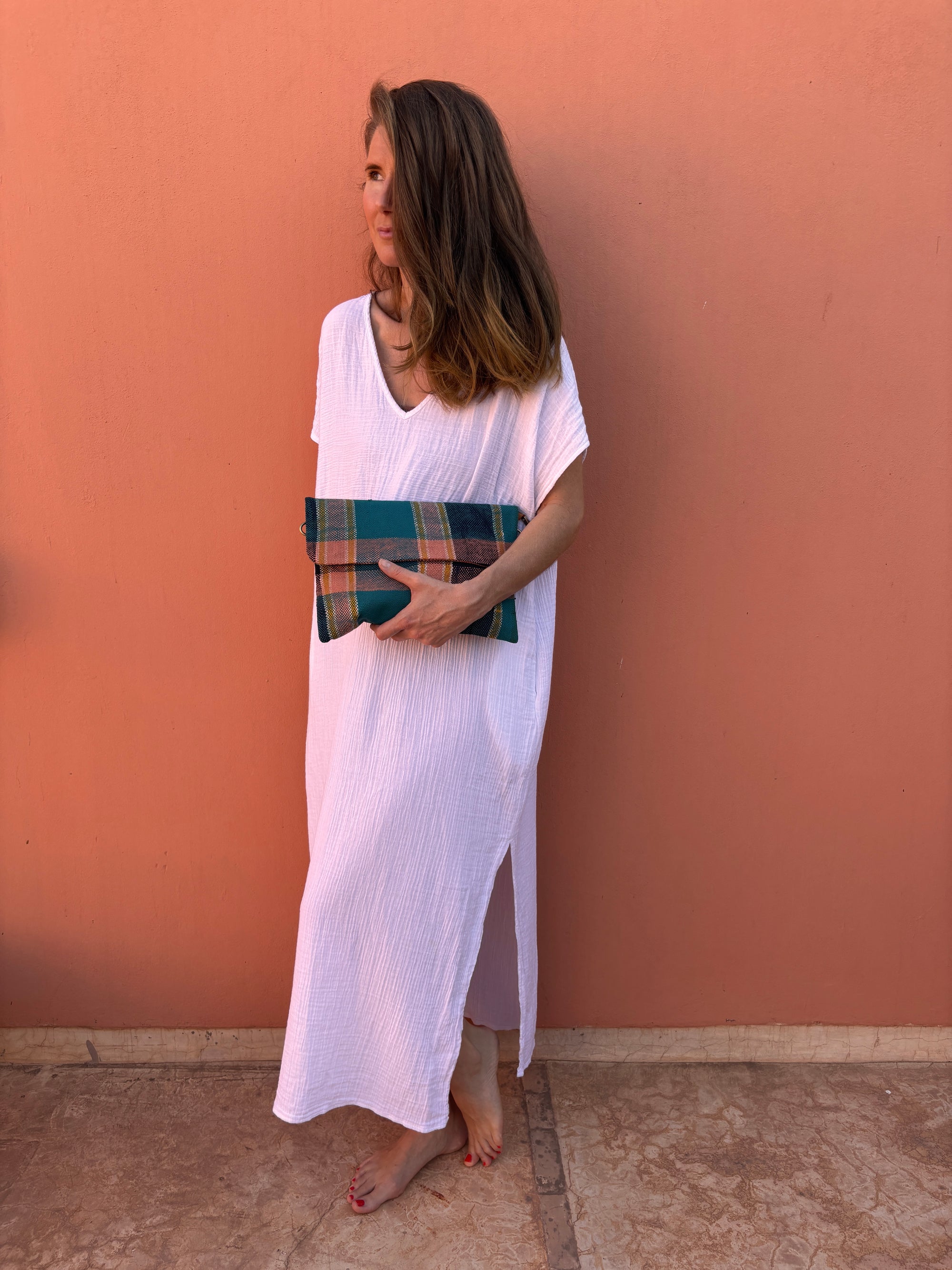 Eine Frau steht barfuß vor einer pfirsichfarbenen Wand, trägt ein weites weißes Kleid und hält die LEY! Vintage Clutch in grünem Wollstoff. Sie blickt mit offenem Haar nach links und strahlt eine entspannte Atmosphäre aus.