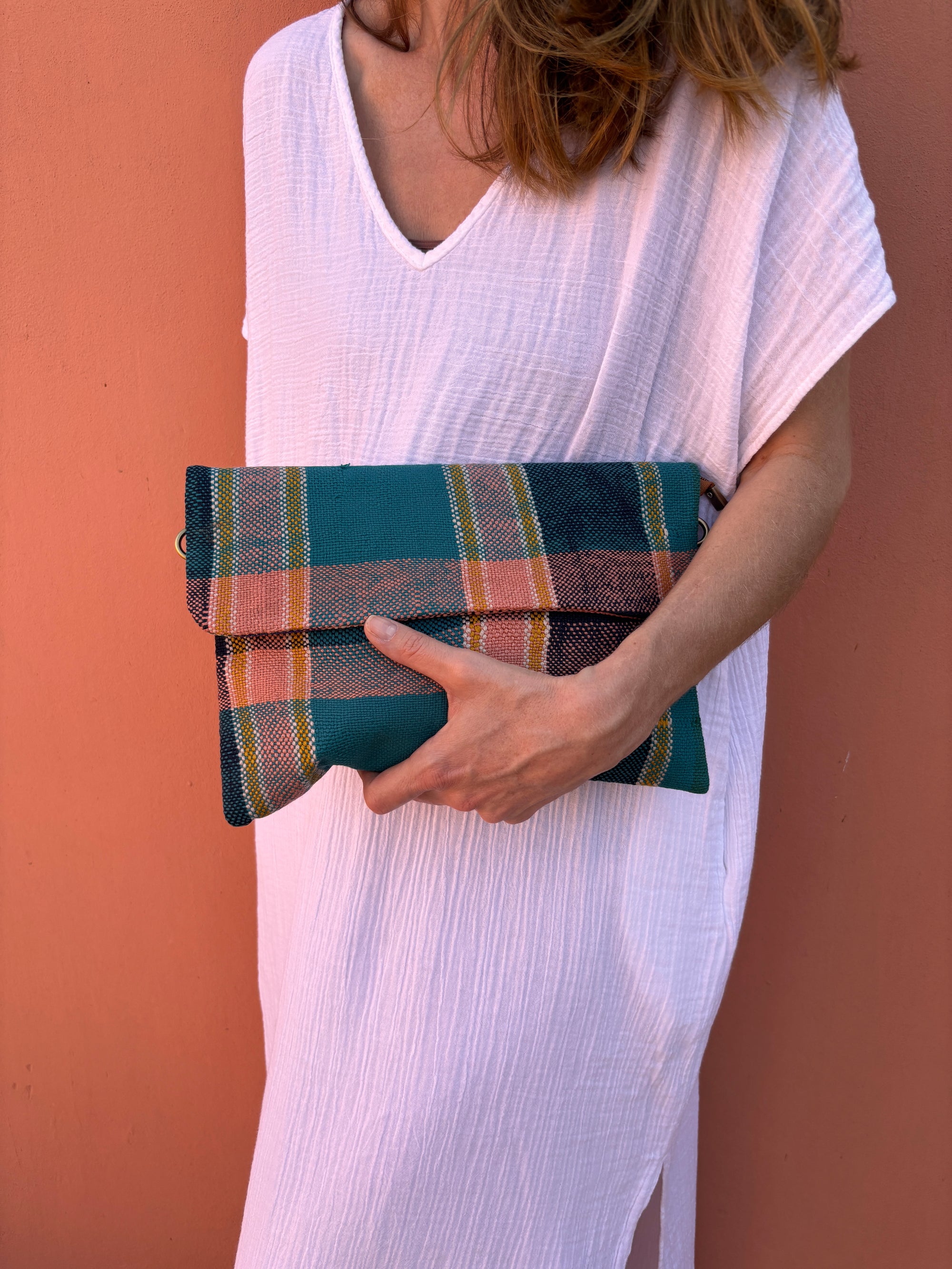 Eine Frau in einem weiten weißen Kleid steht vor einer pfirsichfarbenen Wand und hält die LEY! Vintage Clutch - eine gefaltete rechteckige Tasche mit einem grün, rosa und gelb karierten Muster. Ihr Gesicht bleibt unsichtbar.