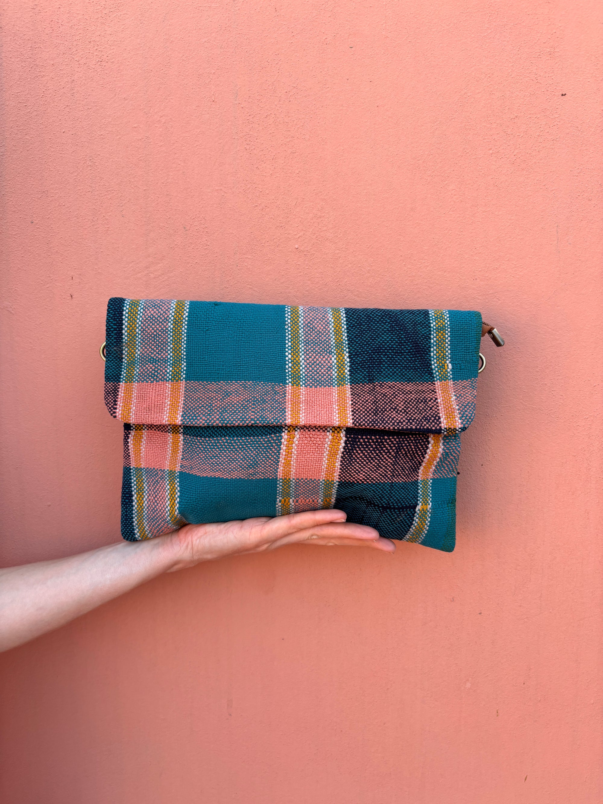 Eine Hand hält die LEY! Vintage Clutch - eine gefaltete, rechteckige Tasche aus Wollstoff mit einem rosa, blau und gelb karierten Muster - vor einer pfirsichfarbenen Wand.