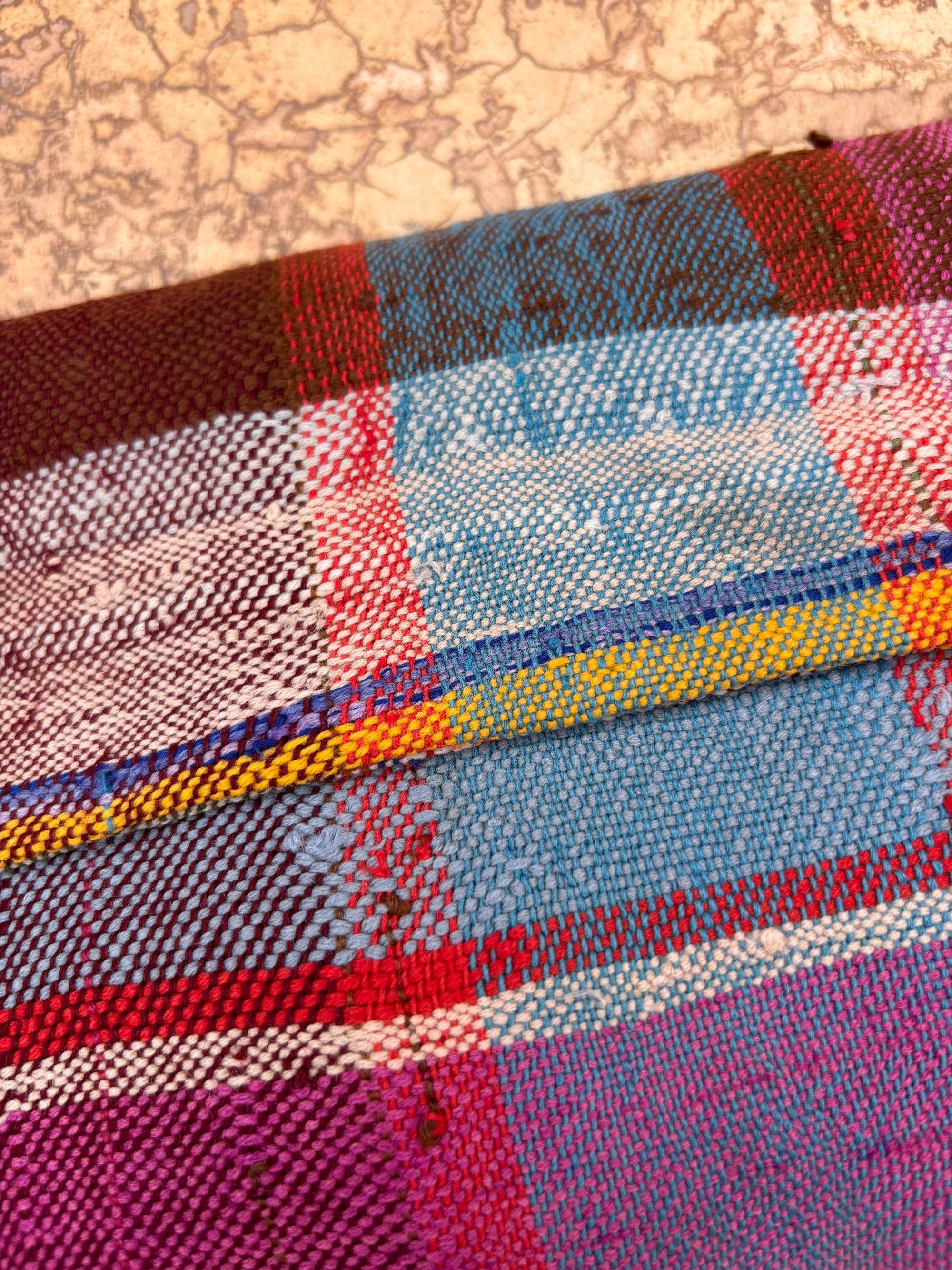 Nahaufnahme eines farbenfrohen gewebten Stoffes aus einem Upcycling-Projekt mit roten, blauen, gelben und lila karierten Fäden. Sichtbare Nähte zeichnen die Naht dieser handgefertigten Vintage Clutch von LEY! mit strukturiertem Hintergrund nach.