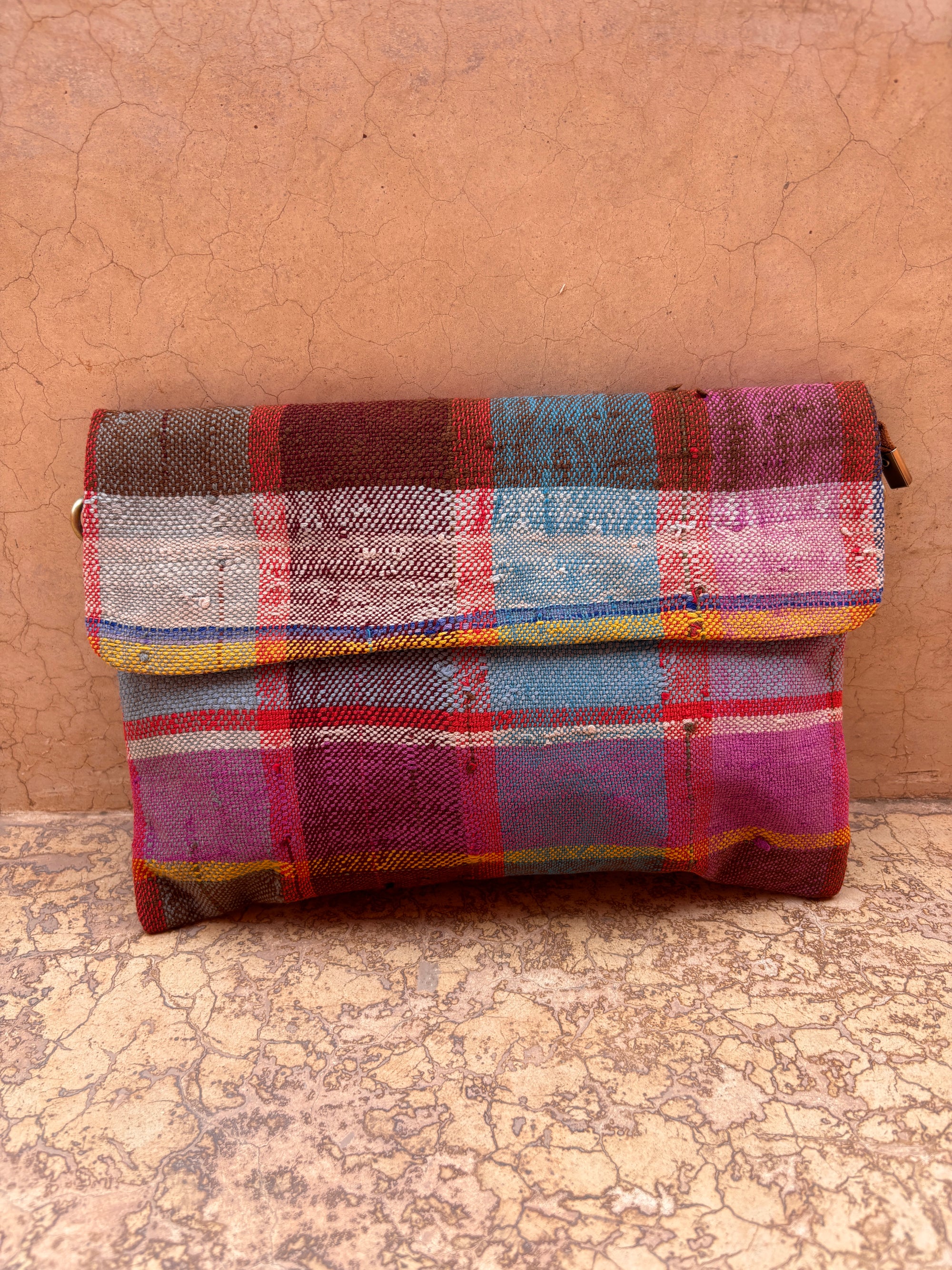 Die LEY! Vintage Clutch ist eine farbenfrohe, rechteckige Tasche mit Klappe, handgefertigt aus karierter Wolle in Rot-, Blau-, Pink- und Gelbtönen und vor einem strukturierten, erdigen Hintergrund.