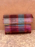 Die LEY! Vintage Clutch ist eine farbenfrohe, rechteckige Tasche mit Klappe, handgefertigt aus karierter Wolle in Rot-, Blau-, Pink- und Gelbtönen und vor einem strukturierten, erdigen Hintergrund.