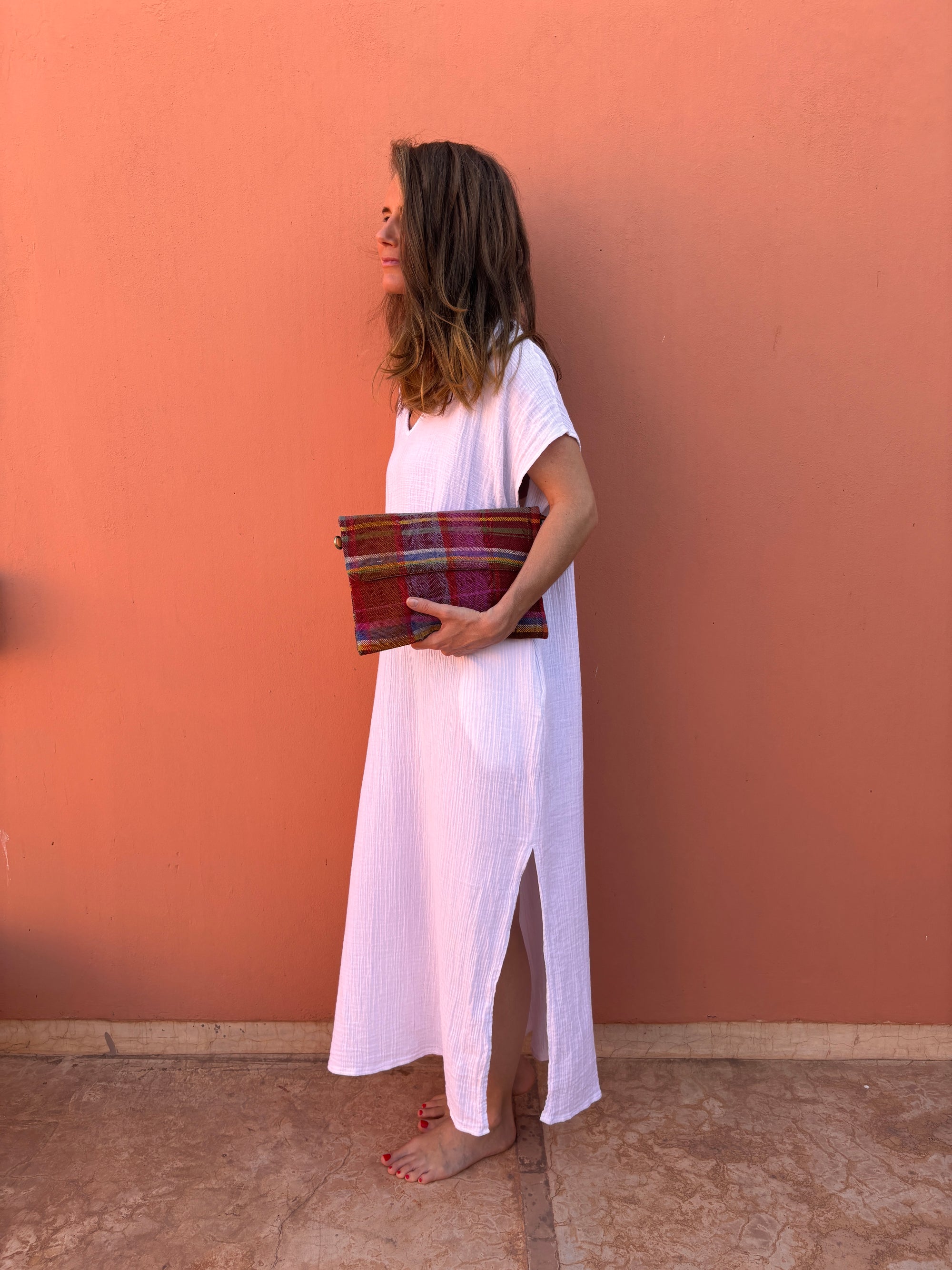 Eine Frau in einem weiten weißen Kleid steht barfuß auf einem Steinboden vor einer pfirsichfarbenen Wand und hält die LEY! Vintage Clutch unter dem Arm und schaut zur Seite.