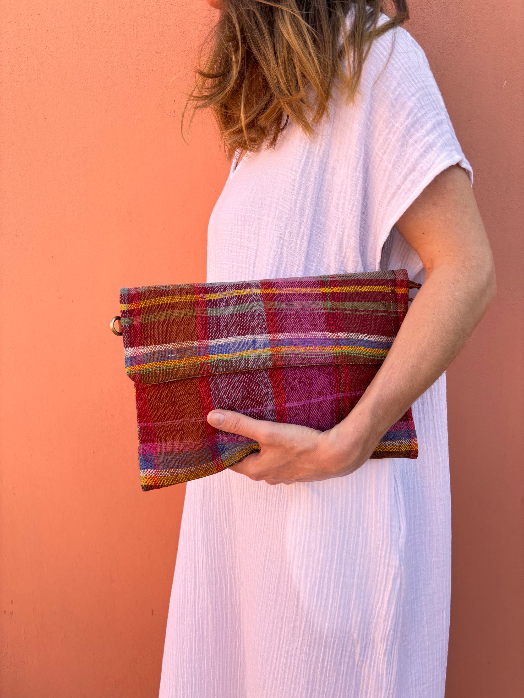 Eine Frau in einem weißen, strukturierten Kleid hält die LEY! Vintage Clutch aus Wolle vor einer pfirsichfarbenen Wand. Ihr Gesicht ist teilweise außerhalb des Rahmens, und ihr gewelltes Haar fällt über ihre Schulter.