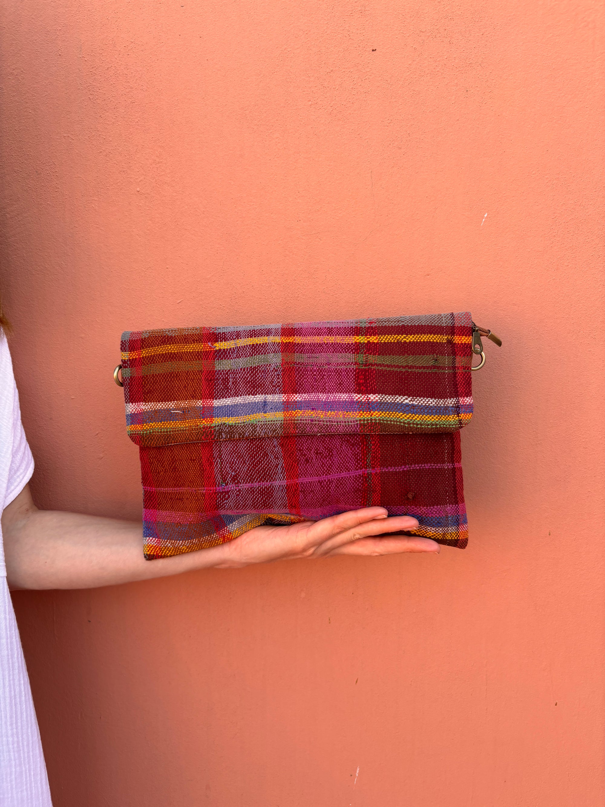 Eine Person in einem weißen Oberteil hält die LEY! Vintage Clutch - eine leuchtend karierte Handtasche in Rot, Pink, Orange und Lila - und hebt ihren Charme vor einer pfirsichfarbenen Wand hervor.
