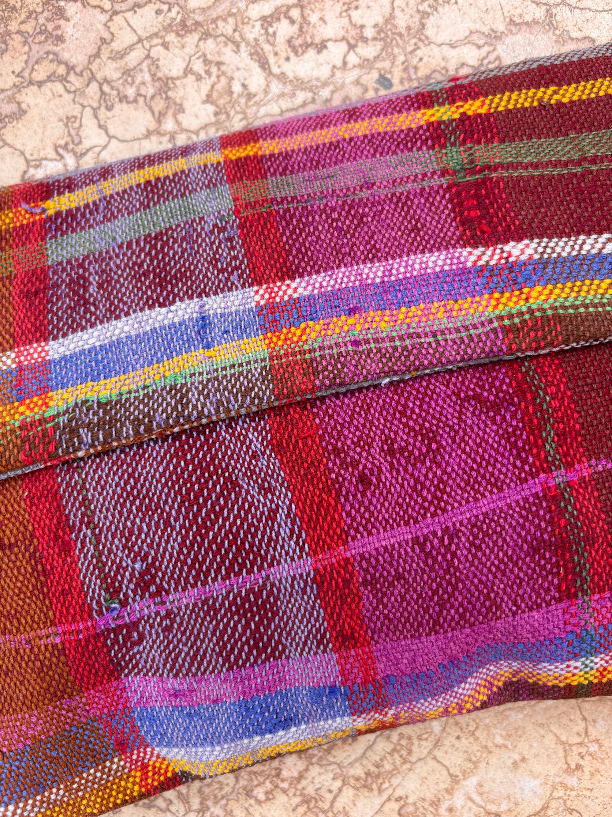 Nahaufnahme eines farbenfrohen, karierten Gewebes in Rot, Lila, Gelb, Blau und Weiß aus der Vintage Clutch von LEY!, das von handgewebten Wolltextilien inspiriert ist und auf einer marmorierten Oberfläche präsentiert wird - perfekt, um Ihrem Look einen einzigartigen Touch zu verleihen.