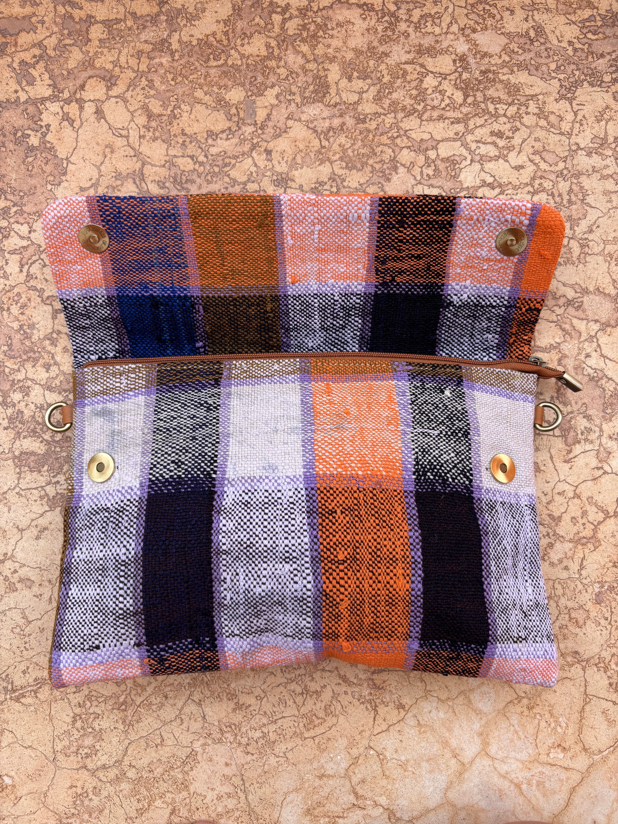 Die LEY! Vintage Clutch ist eine kleine rechteckige Tasche, handgefertigt aus karierter Wolle in orange, schwarz, weiß und lila Streifen. Die Abbildung zeigt sie ohne Reißverschluss auf Stein und mit vier Metallösen an jeder Ecke.