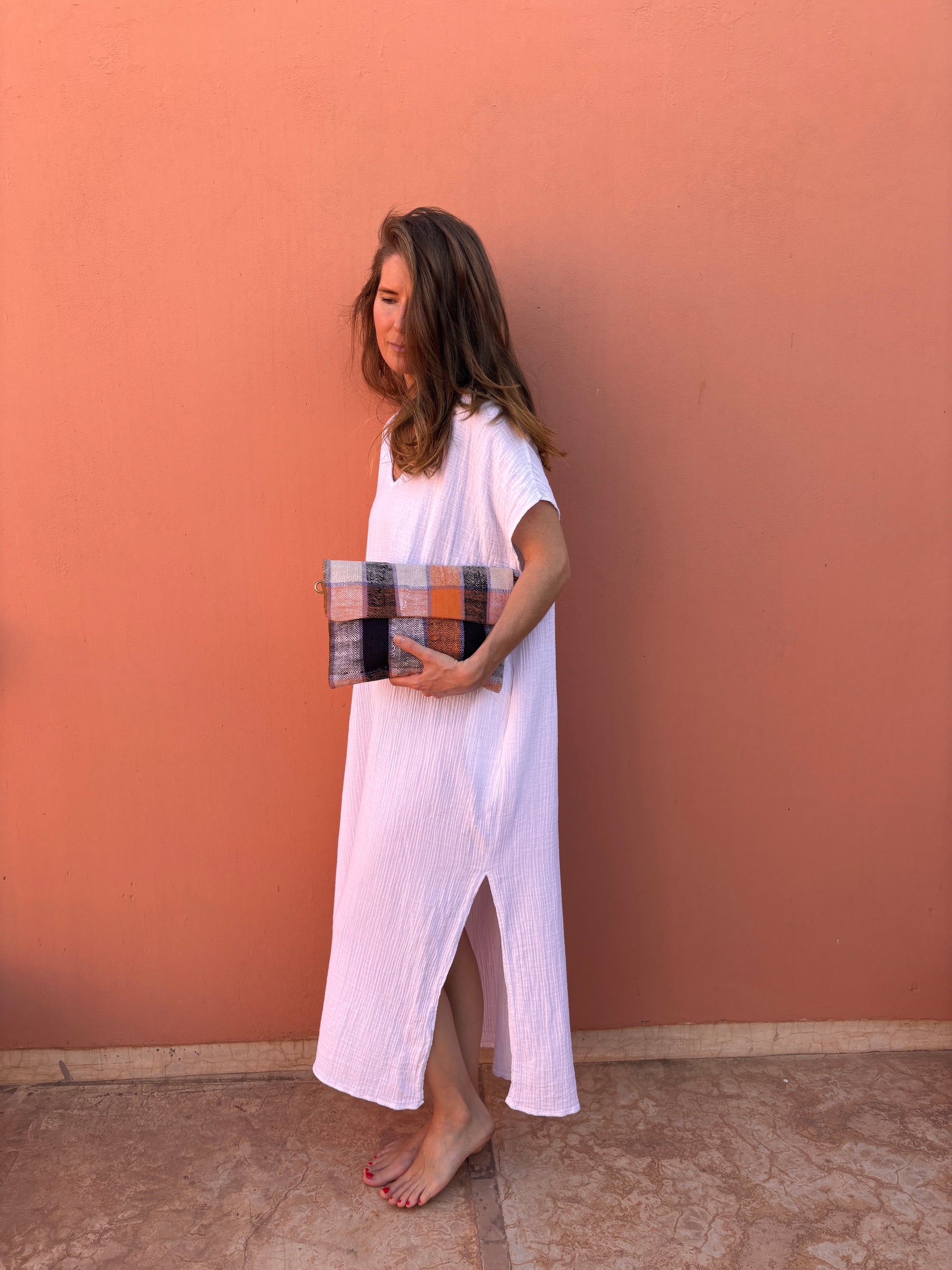 Eine Frau in einem weißen, kurzärmeligen, knöchellangen Kleid steht barfuß auf einem Stein vor einer pfirsichfarbenen Wand und hält die LEY! Vintage Clutch aus orangefarbenen, grauen und violetten Wollstoffen.