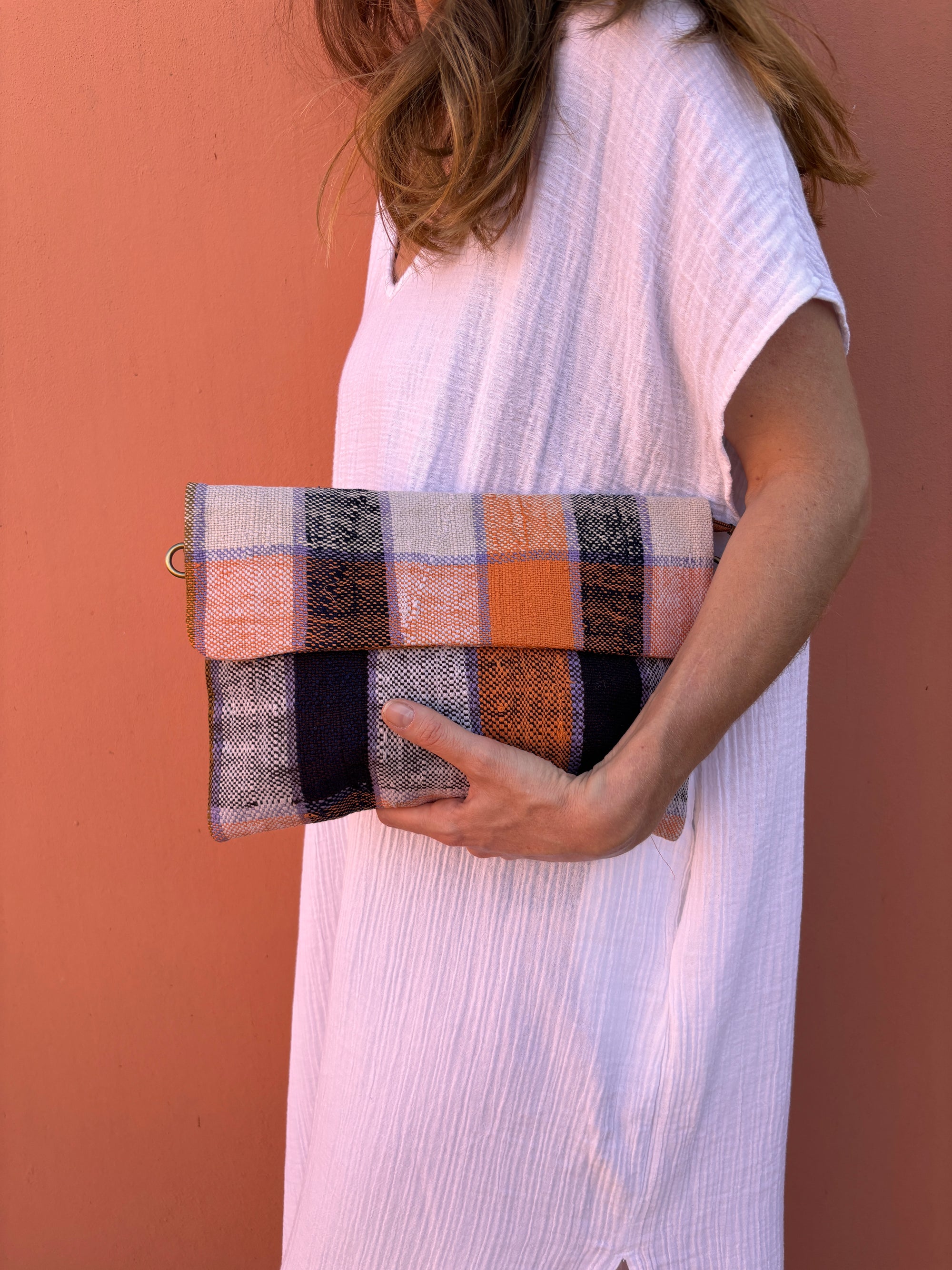 Eine Person in einem lockeren weißen Kleid hält eine LEY! Vintage Clutch in orangefarbenen, marineblauen und cremefarbenen Wollstoffen, die vor einer pfirsichfarbenen Wand steht.