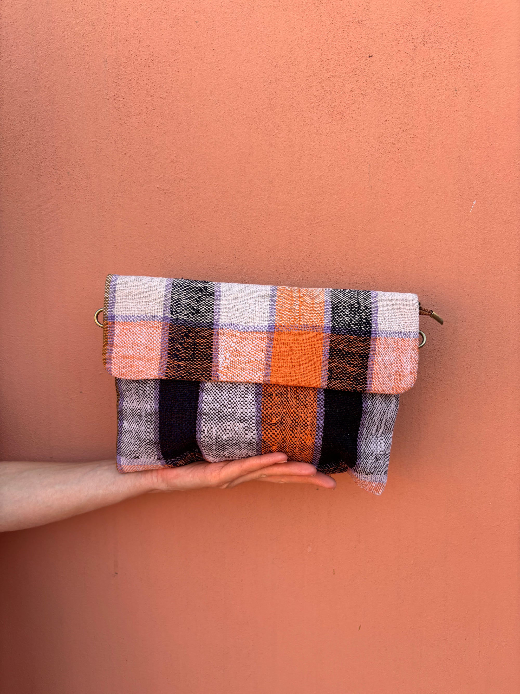 Eine Hand hält die LEY! Vintage Clutch, ein handgefertigtes Stück mit einer Klappe und einem karierten Muster aus orangefarbenem, schwarzem, grauem und weißem Wollstoff vor einem pfirsichfarbenen Hintergrund.