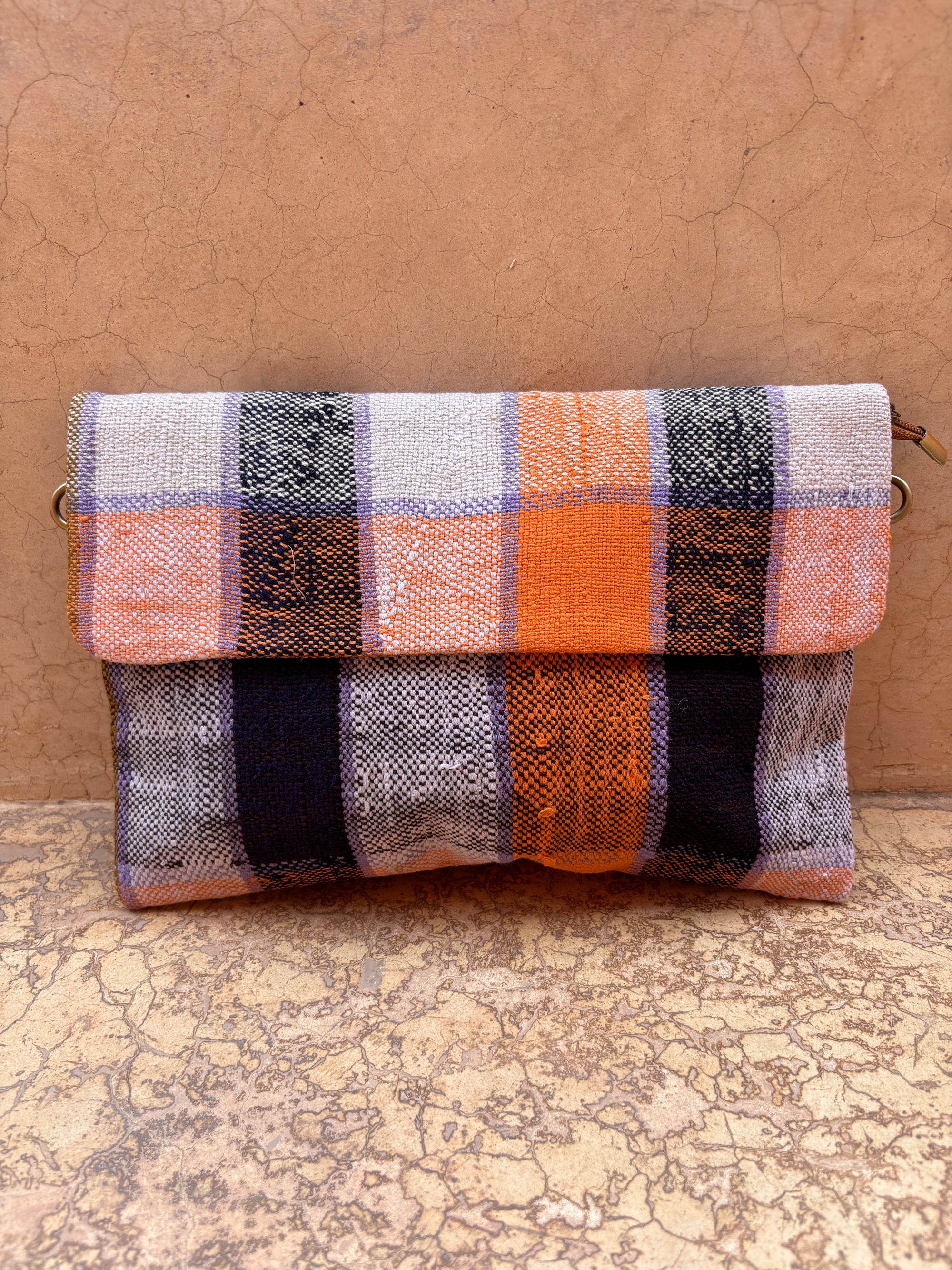 Die LEY! Vintage Clutch aus upgecycelter Wolle ist mit einem gewebten orange-schwarz-grau-weißen Karomuster versehen und steht aufrecht vor einer strukturierten hellbraunen Wand.
