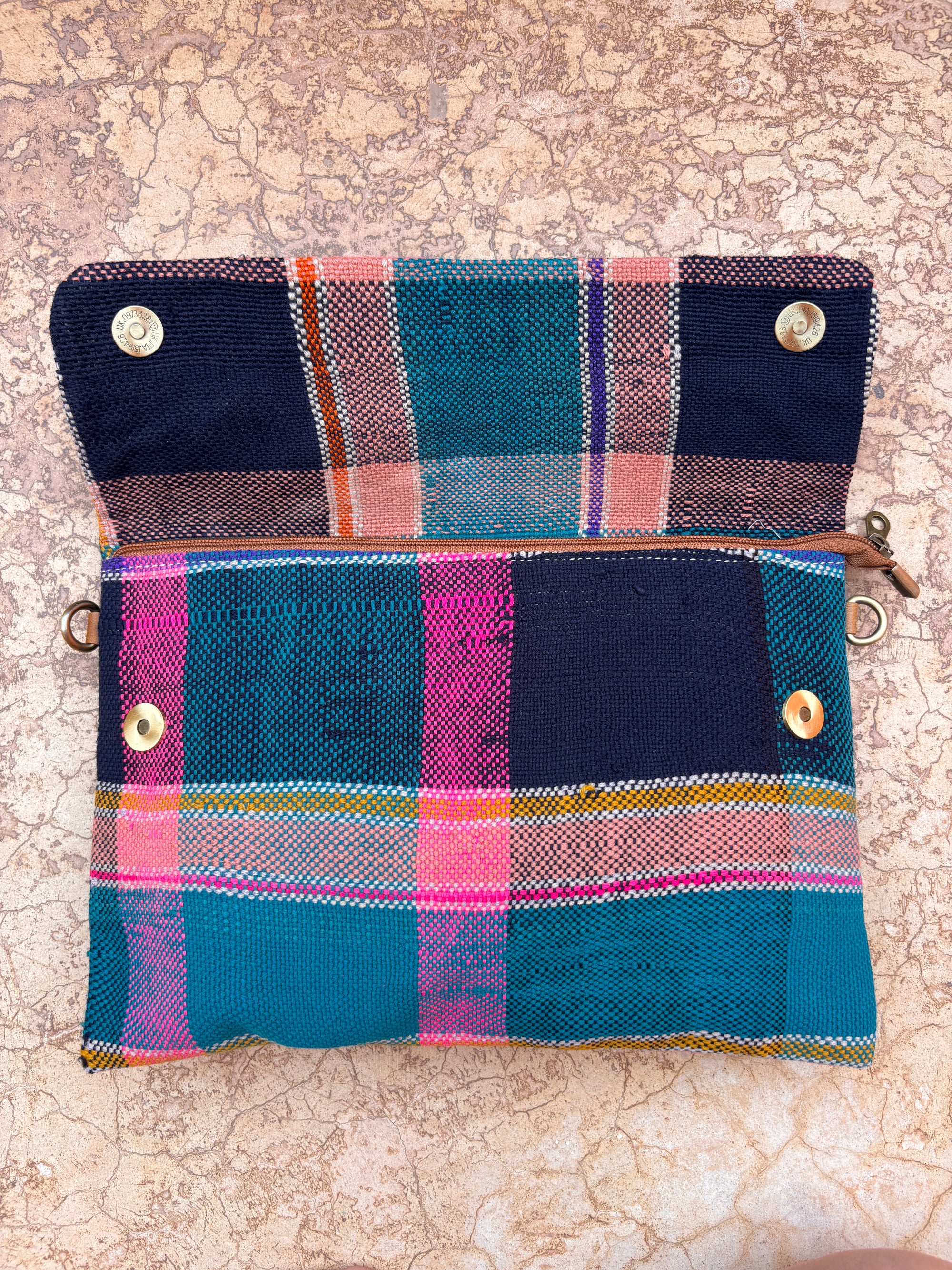Die Vintage Clutch von LEY! zeigt ein farbenfrohes Karomuster mit blauen, rosa, orangen und blaugrünen Streifen in einem gewebten Muster. Diese handgefertigte Tasche hat einen Reißverschluss, Druckknöpfe und wird offen auf einer strukturierten Steinoberfläche gezeigt.