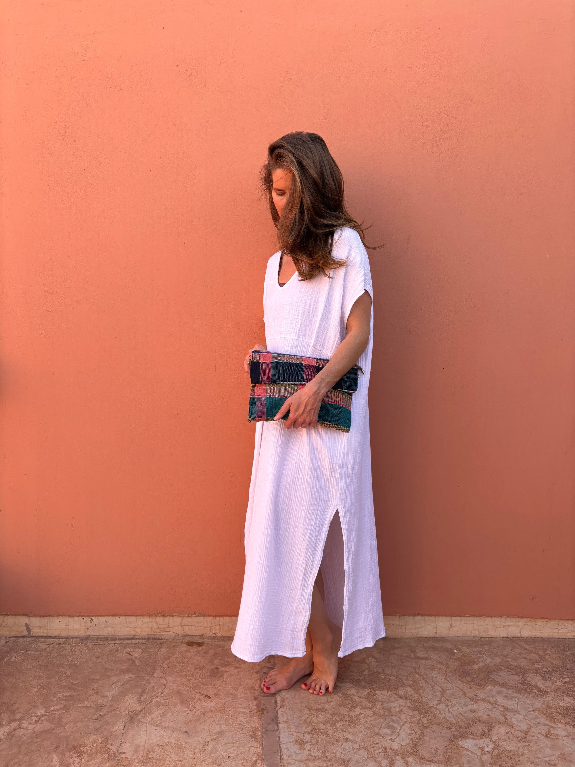 Eine Person steht barfuß vor einer pfirsichfarbenen Wand, trägt ein lockeres weißes, knöchellanges Kleid und hält die LEY! Vintage Clutch. Ihr Haar fällt locker und verdeckt teilweise ihr Gesicht, während sie leicht nach unten schaut.