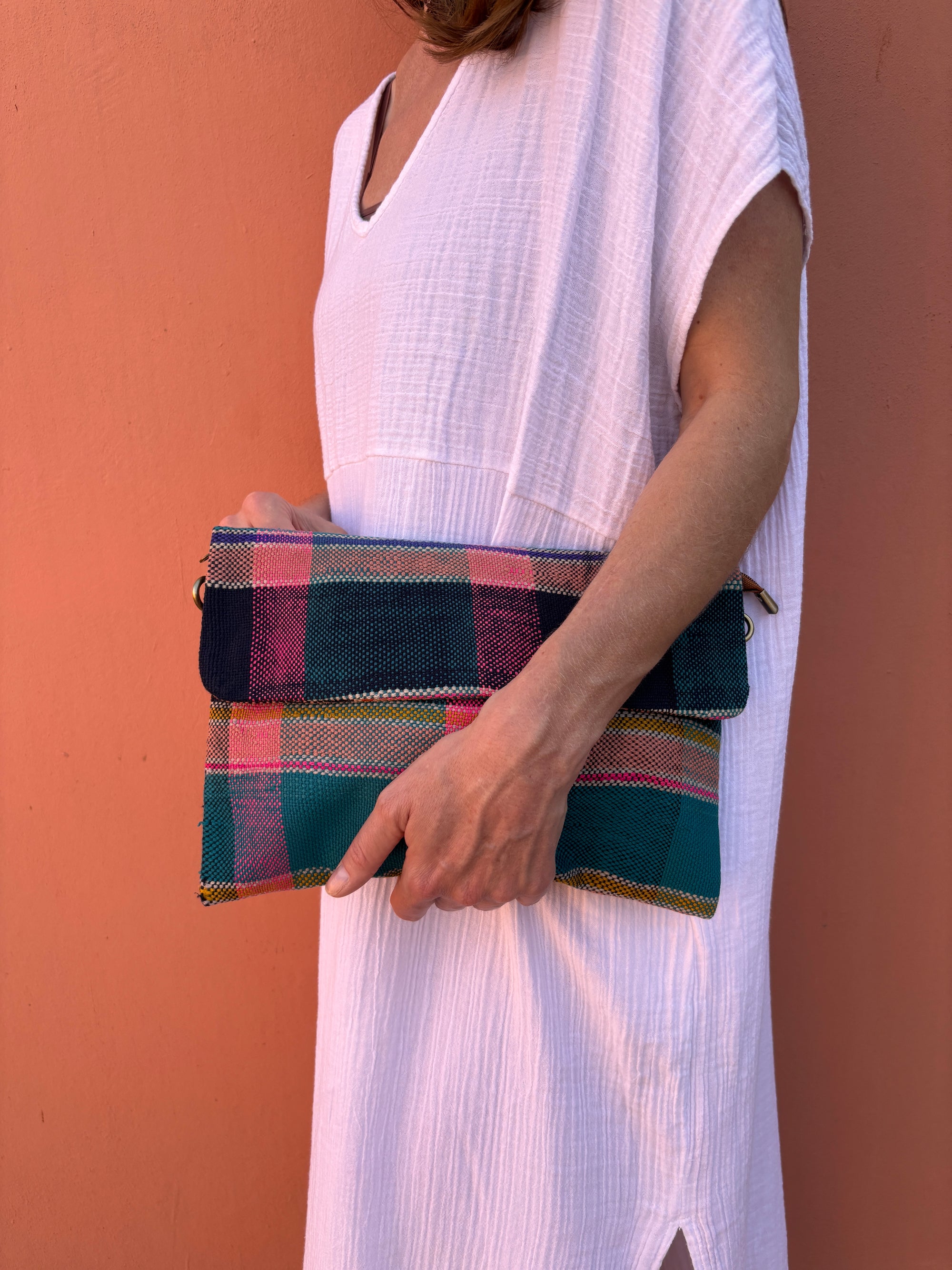 Eine Person in einem weißen kurzärmeligen Kleid hält die LEY! Vintage Clutch, eine bunte, handgefertigte Wolltasche, gegen eine pfirsichfarbene Wand. Nur der Oberkörper und ein Teil eines Arms sind zu sehen.