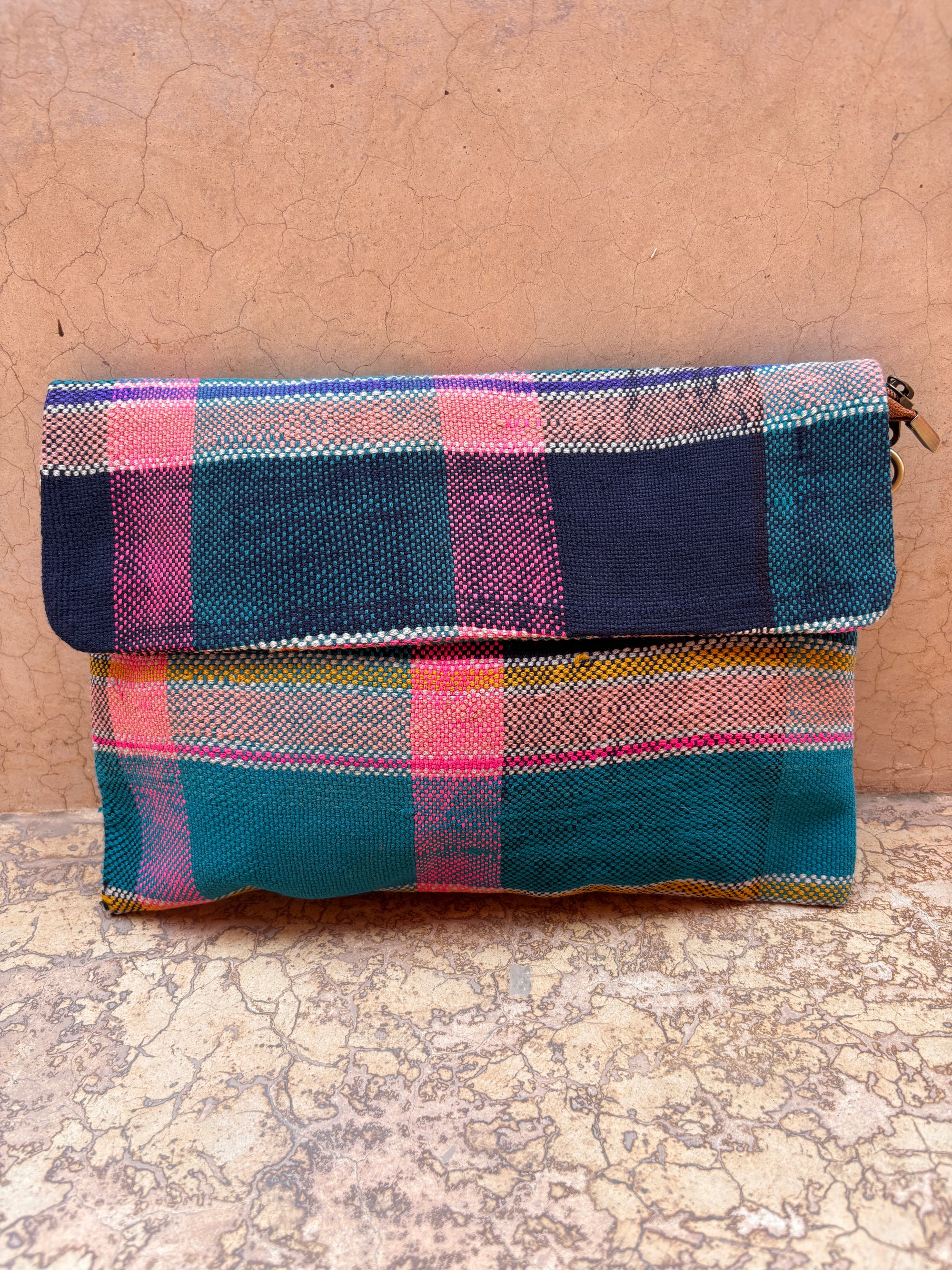 Die Vintage Clutch von LEY! zeigt ein leuchtendes Karomuster in Pink, Blau und Petrol und ruht auf einer strukturierten Steinoberfläche mit passendem Hintergrund - ein herausragendes Accessoire für jede Sammlung.