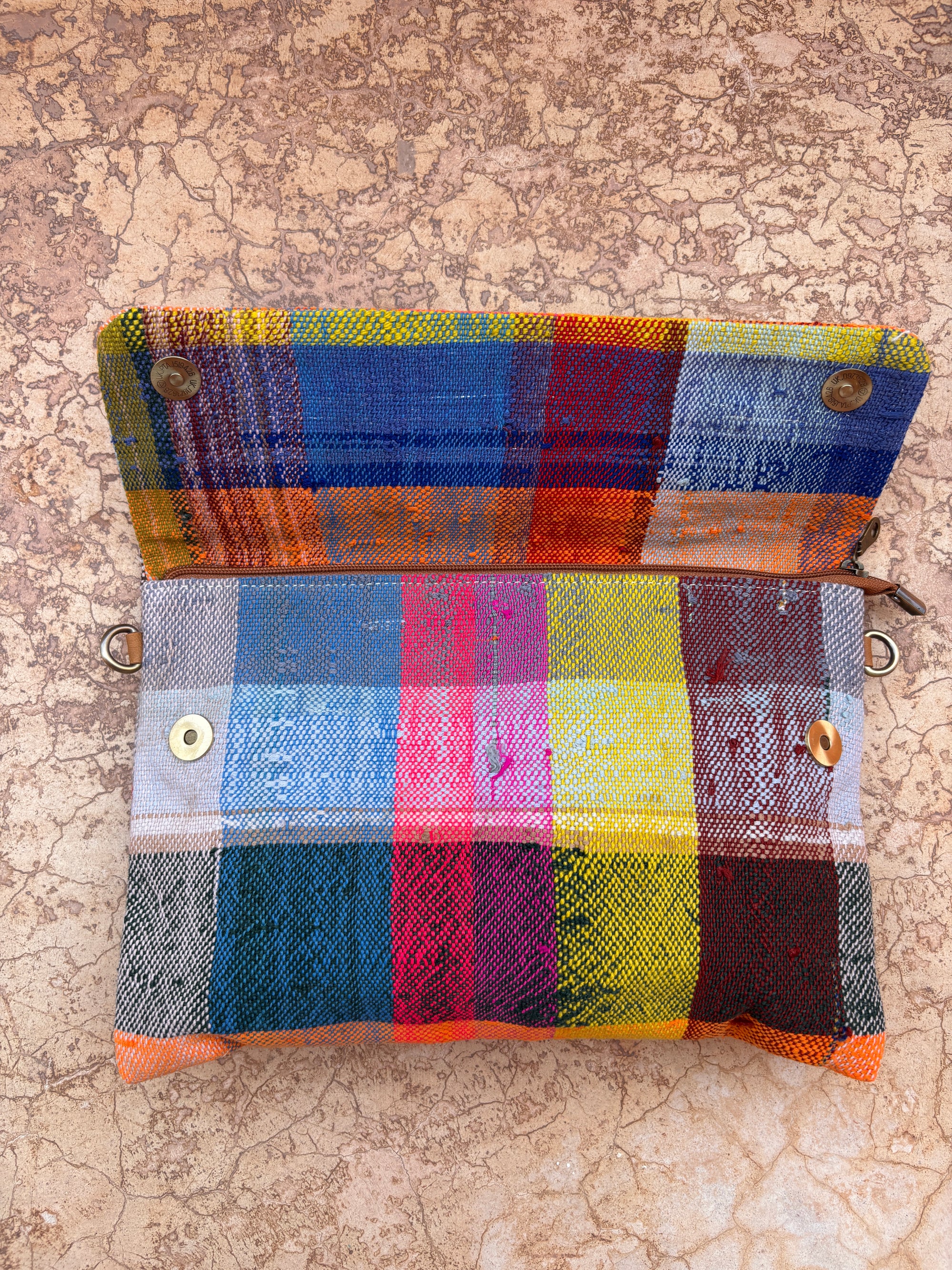 Die LEY! Vintage Clutch, handgefertigt aus Upcycling Wolle, präsentiert ein farbenfrohes Karo-Muster in Rot, Blau, Gelb, Orange und Grün. Mit Reißverschluss und Druckknöpfen liegt sie geöffnet auf einer rissigen Steinfläche.