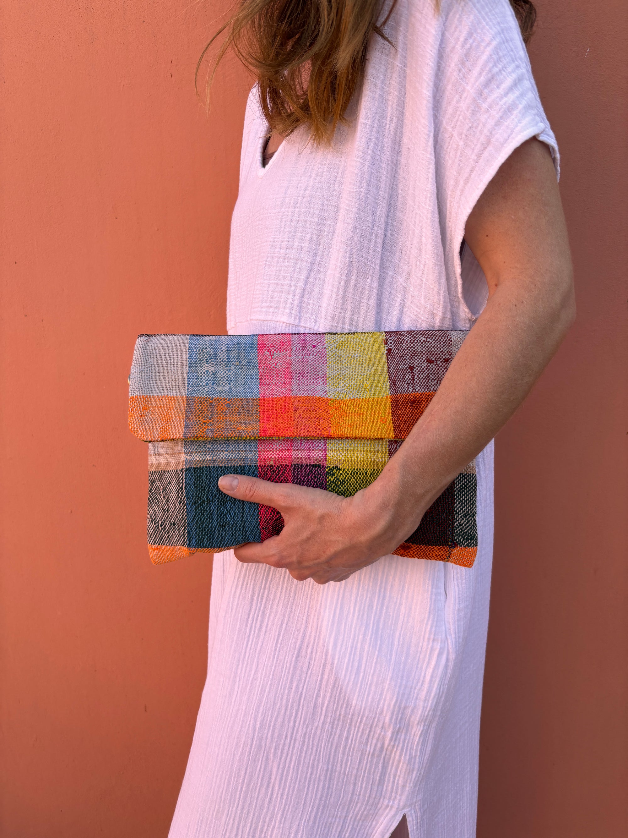 Eine Person in einem kurzärmeligen weißen strukturierten Kleid hält die LEY! Vintage Clutch, die aus upgecycelter Wolle hergestellt wurde, vor einer pfirsichfarbenen Wand. Nur ihr Oberkörper und ihr Arm sind zu sehen.