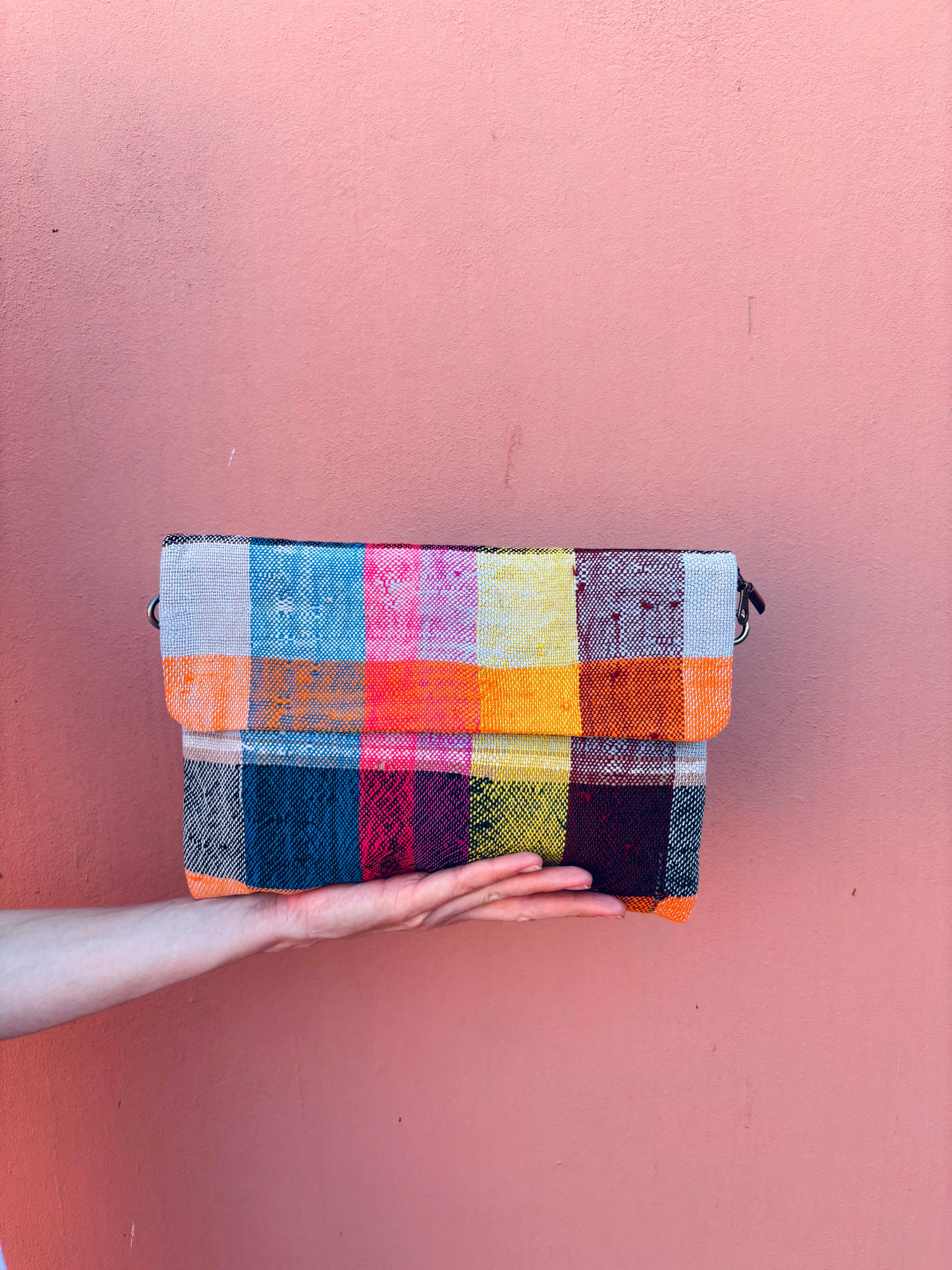 Eine Hand hält die LEY! Vintage Clutch, eine handgefertigte rechteckige Tasche mit einem farbenfrohen Karomuster in Blau, Rosa, Gelb, Orange und Weiß - hergestellt aus upgecycelter Wolle - vor einem einfarbigen rosa Hintergrund.