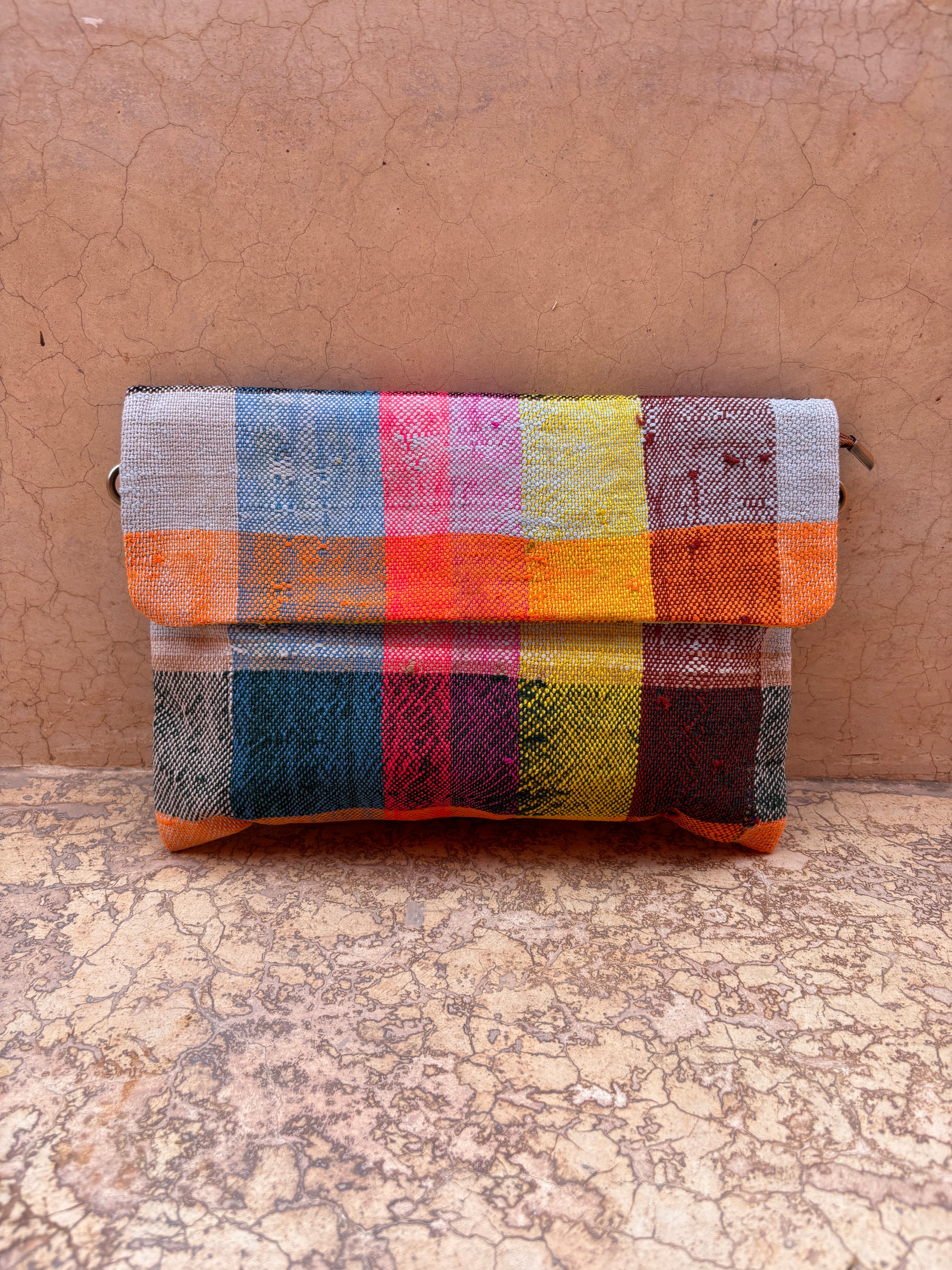 Die LEY! Vintage Clutch ist eine handgefertigte, mehrfarbige Tasche mit einem rosa, gelben, orangen, blauen, schwarzen und grauen karierten Upcycling-Wolle-Design vor einem strukturierten Wand- und Bodenhintergrund.