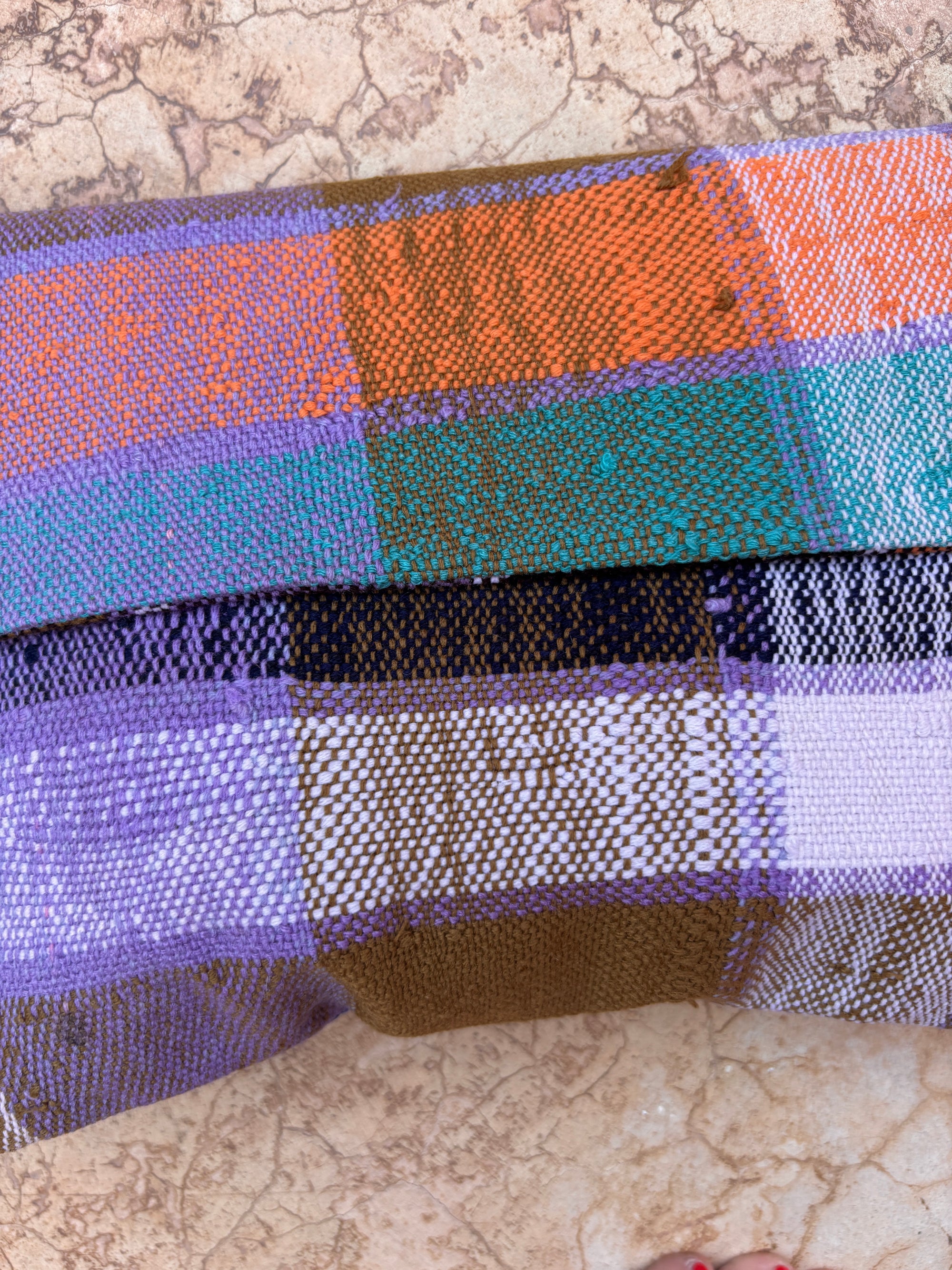 Eine Nahaufnahme einer farbenfrohen gewebten Decke mit orangefarbenen, grünen, lila, braunen und weißen Streifen ruht auf einem strukturierten beigen Stein - die perfekte Inspiration für eine Vintage Clutch von LEY! oder für die Herstellung einer eigenen handgefertigten Tasche.