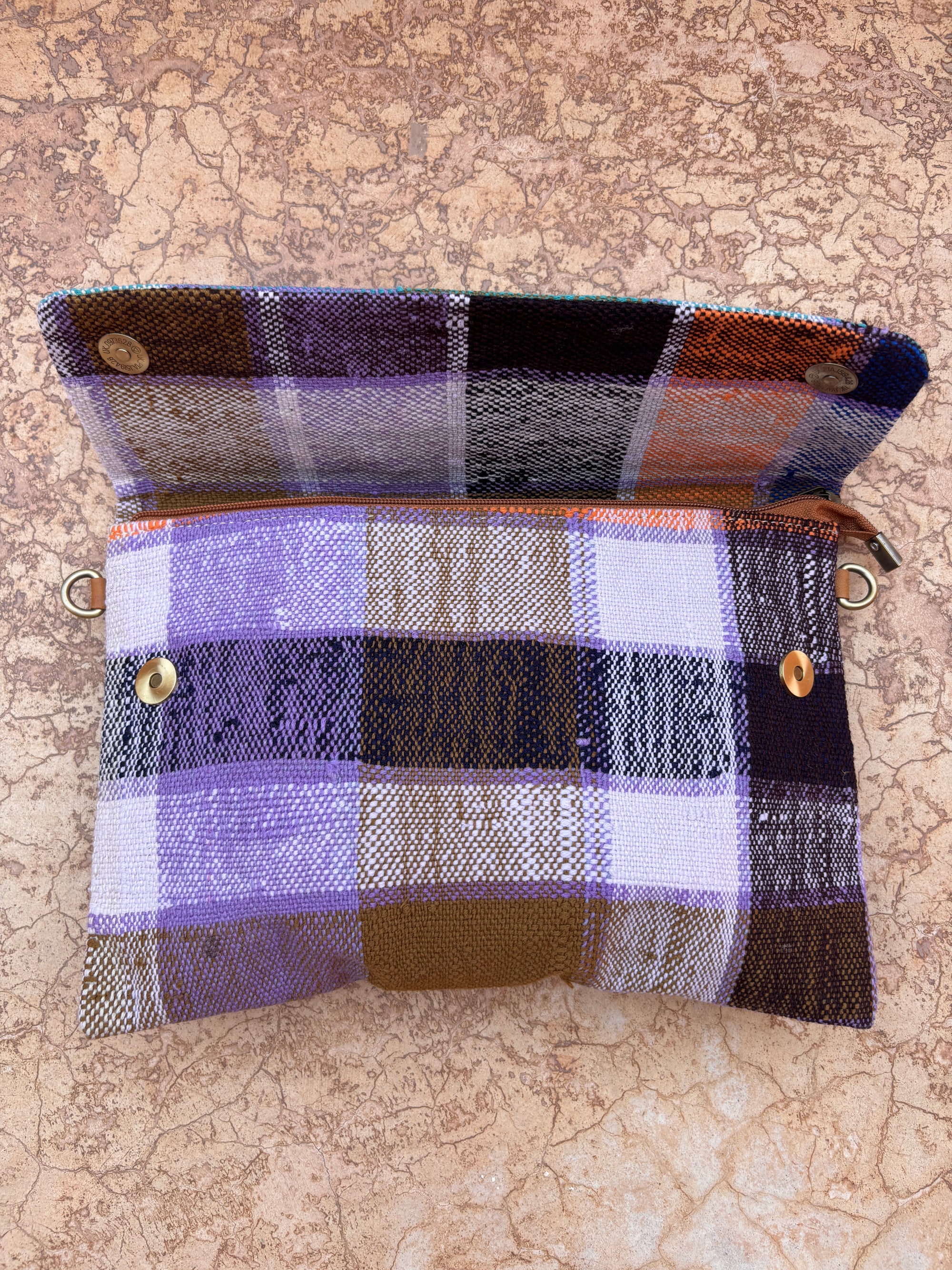 Die LEY! Vintage Clutch ist eine kleine, rechteckige, handgefertigte Tasche mit einem lila, schwarzen, braunen und weißen Karomuster mit einem braunen Reißverschluss und zwei Druckknöpfen, die offen auf einer strukturierten beigen Steinoberfläche gezeigt wird.