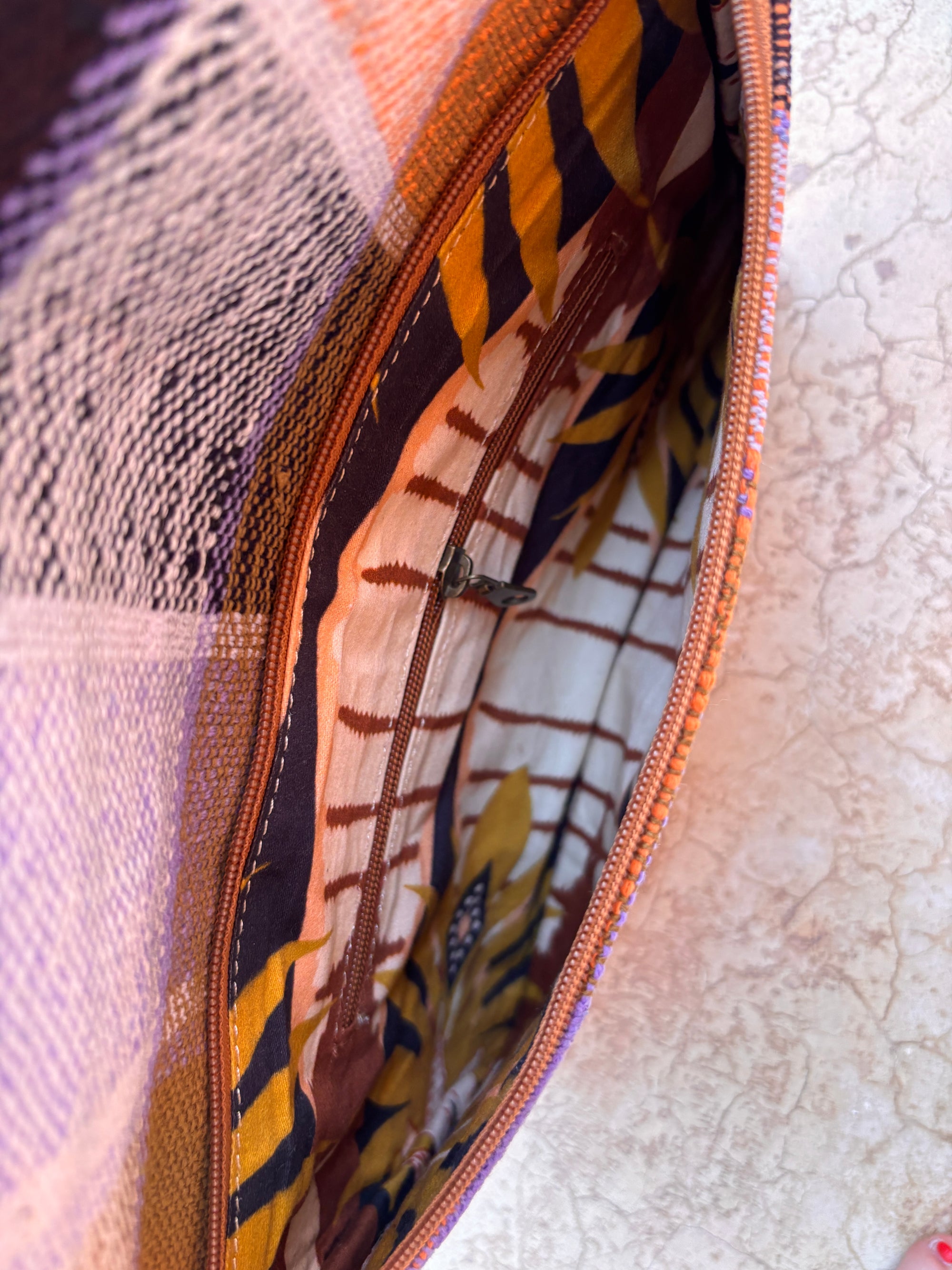 Eine Nahaufnahme der LEY! Vintage Clutch zeigt das handgefertigte Design und das lebendige, gemusterte Futter in Gelb, Orange, Schwarz und Weiß. Dieses upgecycelte Accessoire ruht auf einer leicht strukturierten Oberfläche.