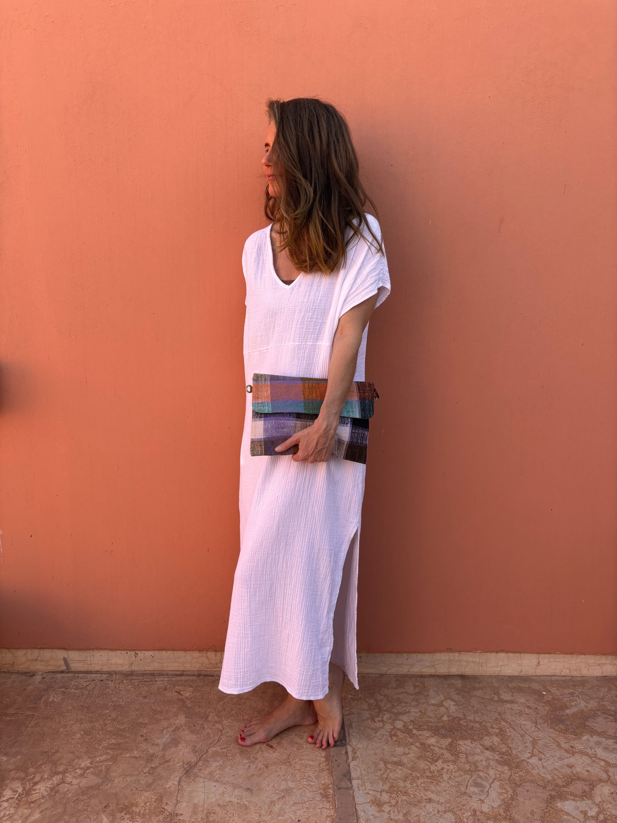 Eine Frau in einem weiten weißen Kleid steht barfuß vor einer pfirsichfarbenen Wand, hält die LEY! Vintage Clutch und schaut nach links.