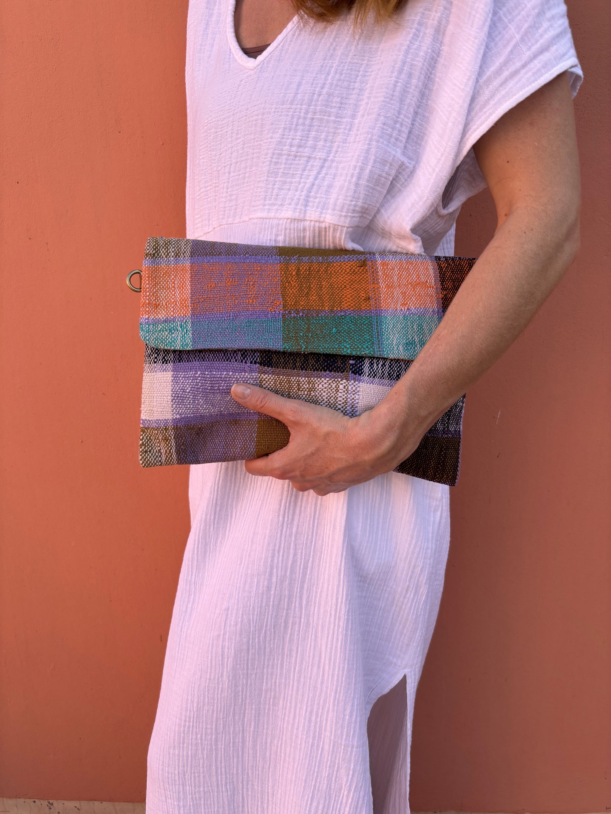 Eine Person in einem weißen, strukturierten Kleid hält die LEY! Vintage Clutch, ein bunt gestreiftes Accessoire, gegen eine pfirsichfarbene Wand. Nur ihr unteres Gesicht und ihr Oberkörper sind sichtbar.