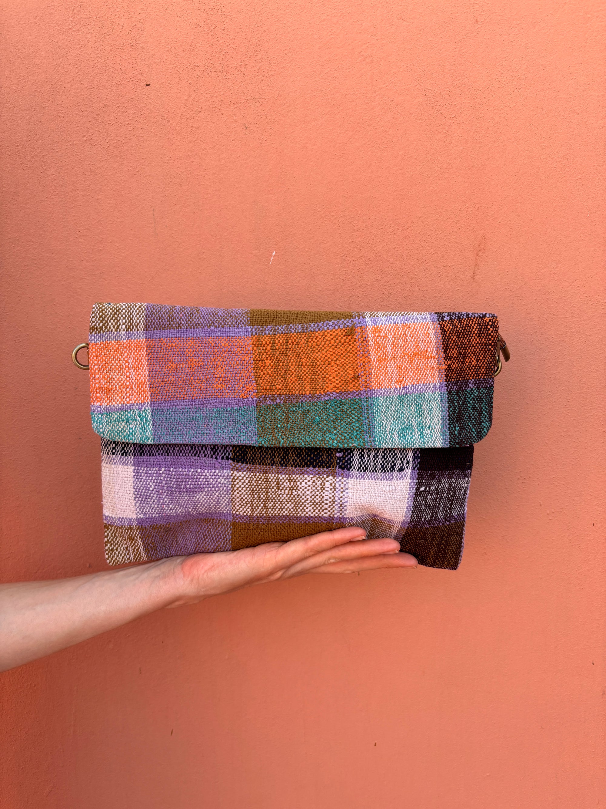 Eine Hand hält die LEY! Vintage Clutch, ein handgefertigtes Accessoire mit einem karierten Muster in Orange, Lila, Grün, Weiß und Braun, vor einer pfirsichfarbenen Wand.