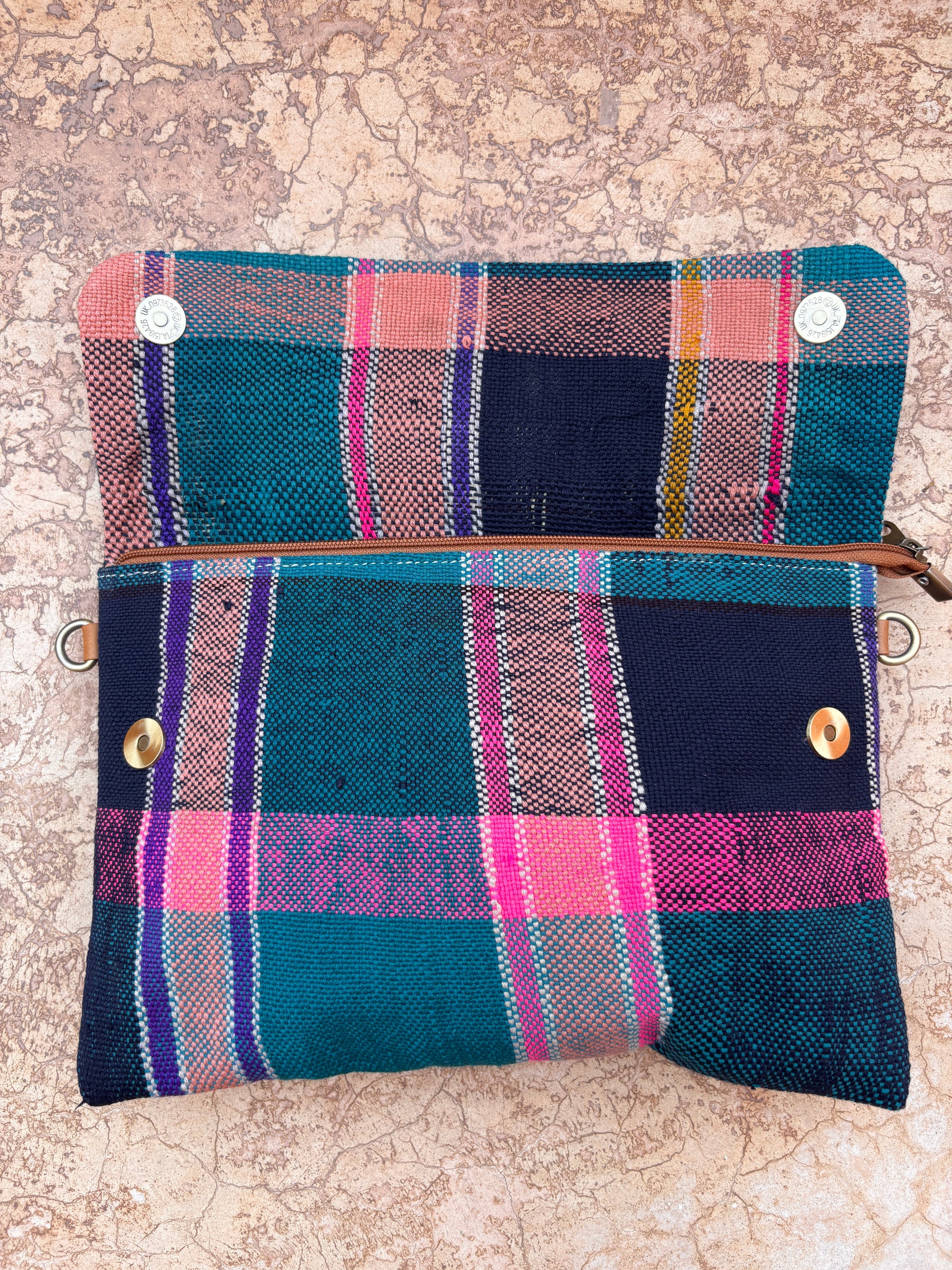 Die LEY! Vintage Clutch mit einem bunten Karomuster mit blauen, blaugrünen, rosa und gelben Streifen liegt offen auf einer strukturierten beigen Oberfläche. Sie ist handgefertigt mit einem Reißverschluss und Druckknöpfen für zusätzlichen Charme.