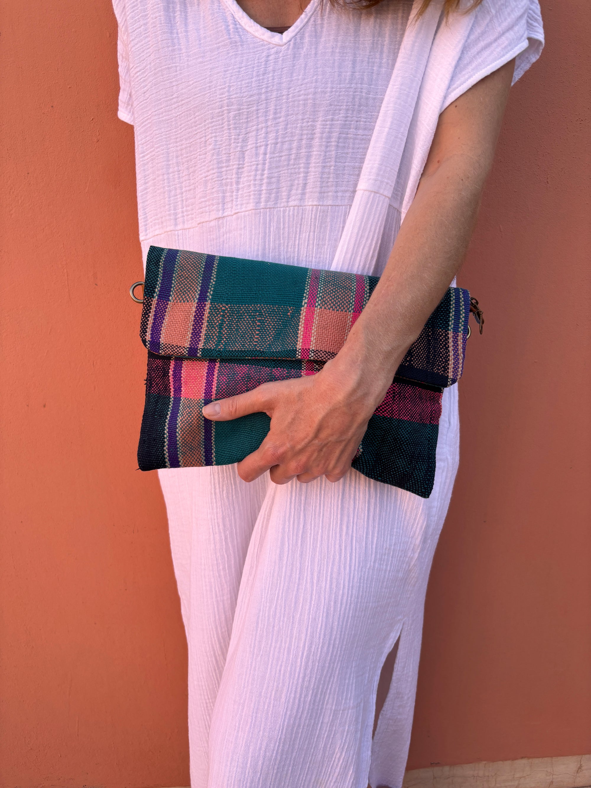 Eine Person in einem weißen, strukturierten Kleid steht vor einer pfirsichfarbenen Wand und hält die LEY! Vintage Clutch aus bunt karierter Wolle in Petrol, Pink und Schwarz. Nur ihr unteres Gesicht, ihr Oberkörper und ihr Arm sind sichtbar.