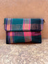 Die LEY! Vintage Clutch ist aus Wolle mit einem Karomuster in Petrol, Rosa, Lila und Schwarz gefertigt und ruht auf einem strukturierten, gebrochenen hellbraunen Hintergrund.