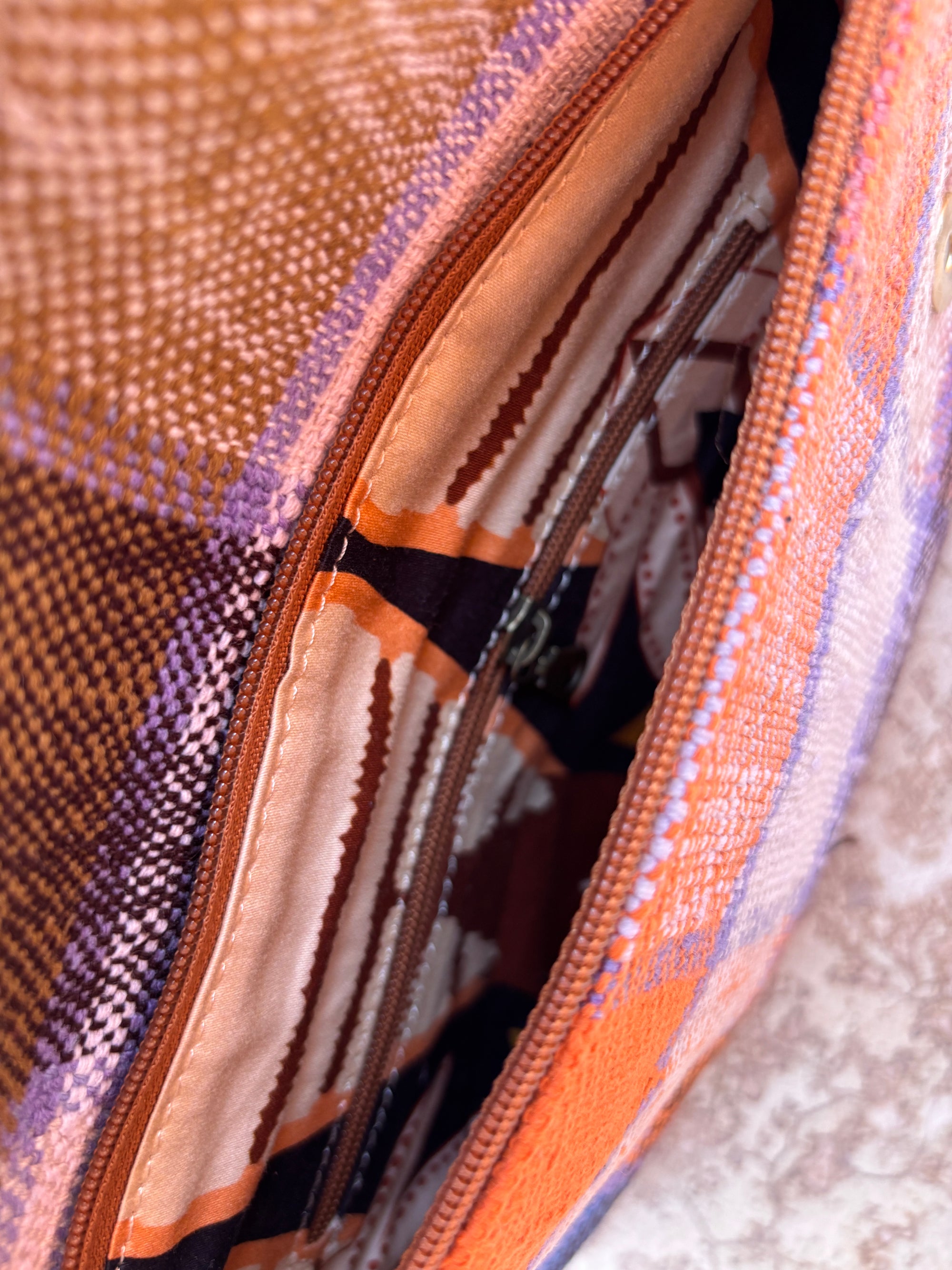 Nahaufnahme einer LEY! Vintage Clutch, die ihr Stofffutter und die Innentasche mit Reißverschluss zeigt. Die Außenseite zeigt ein kariertes Design in Orange-, Lila- und Brauntönen, das aus feiner Wolle gefertigt ist und einen zeitlosen Look bietet.