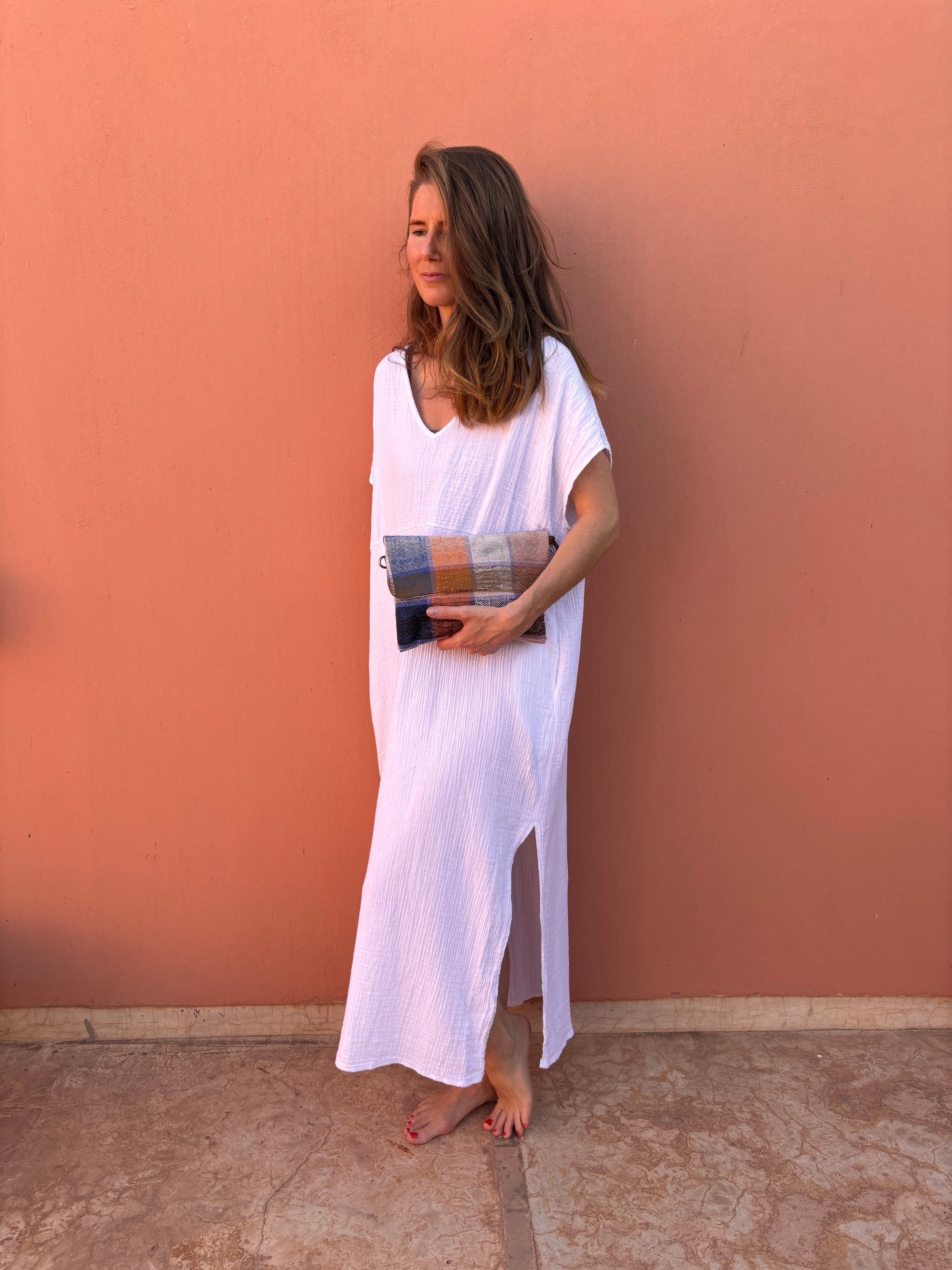 Eine Frau steht barfuß vor einer pfirsichfarbenen Wand, trägt ein weites weißes Kleid und hält die LEY! Vintage Clutch. Ihr Haar ist offen, und sie blickt mit entspanntem Blick zur Seite.