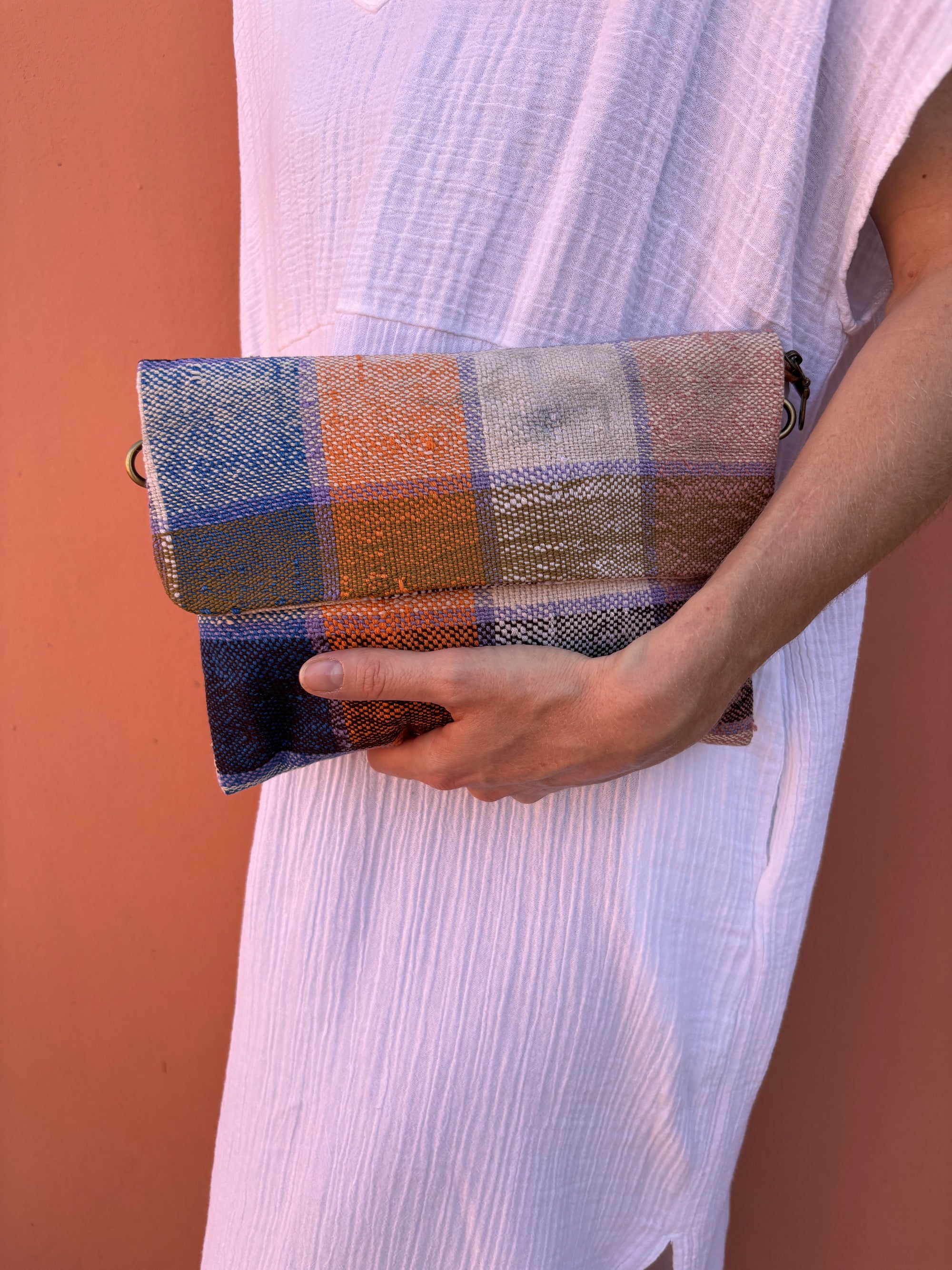 Eine Person in einem weißen, strukturierten Kleid hält die LEY! Vintage Clutch - eine farbenfrohe, handgefertigte Tasche mit einem Karomuster in Orange, Blau, Beige und Braun - während sie vor einer pfirsichfarbenen Wand steht.