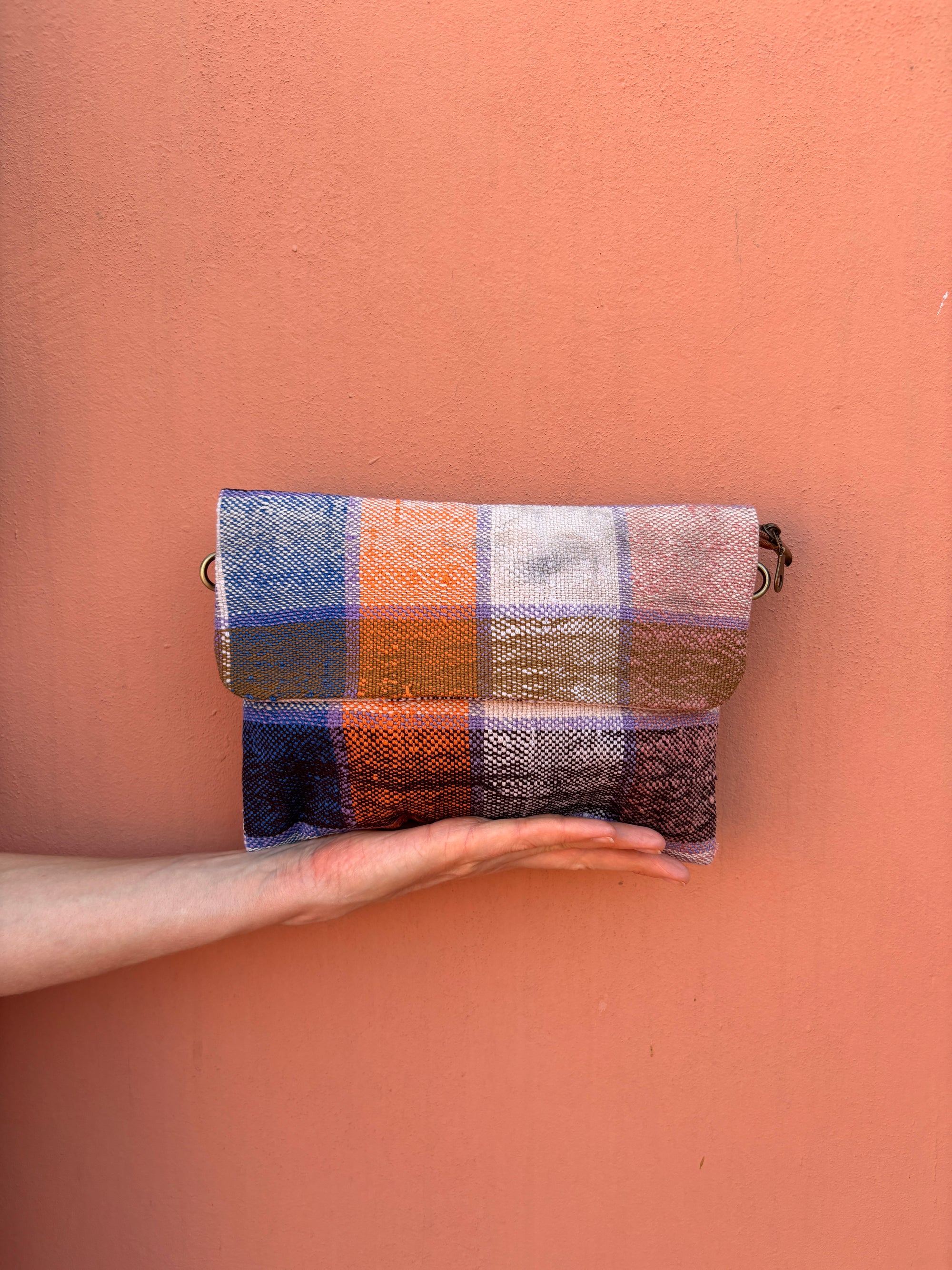 Eine Hand hält die LEY! Vintage Clutch, eine rechteckige, mehrfarbige Tasche mit blauen, orangefarbenen, hellbraunen und weißen vertikalen Streifen - ein auffälliges Accessoire, das zu jedem Anlass passt.