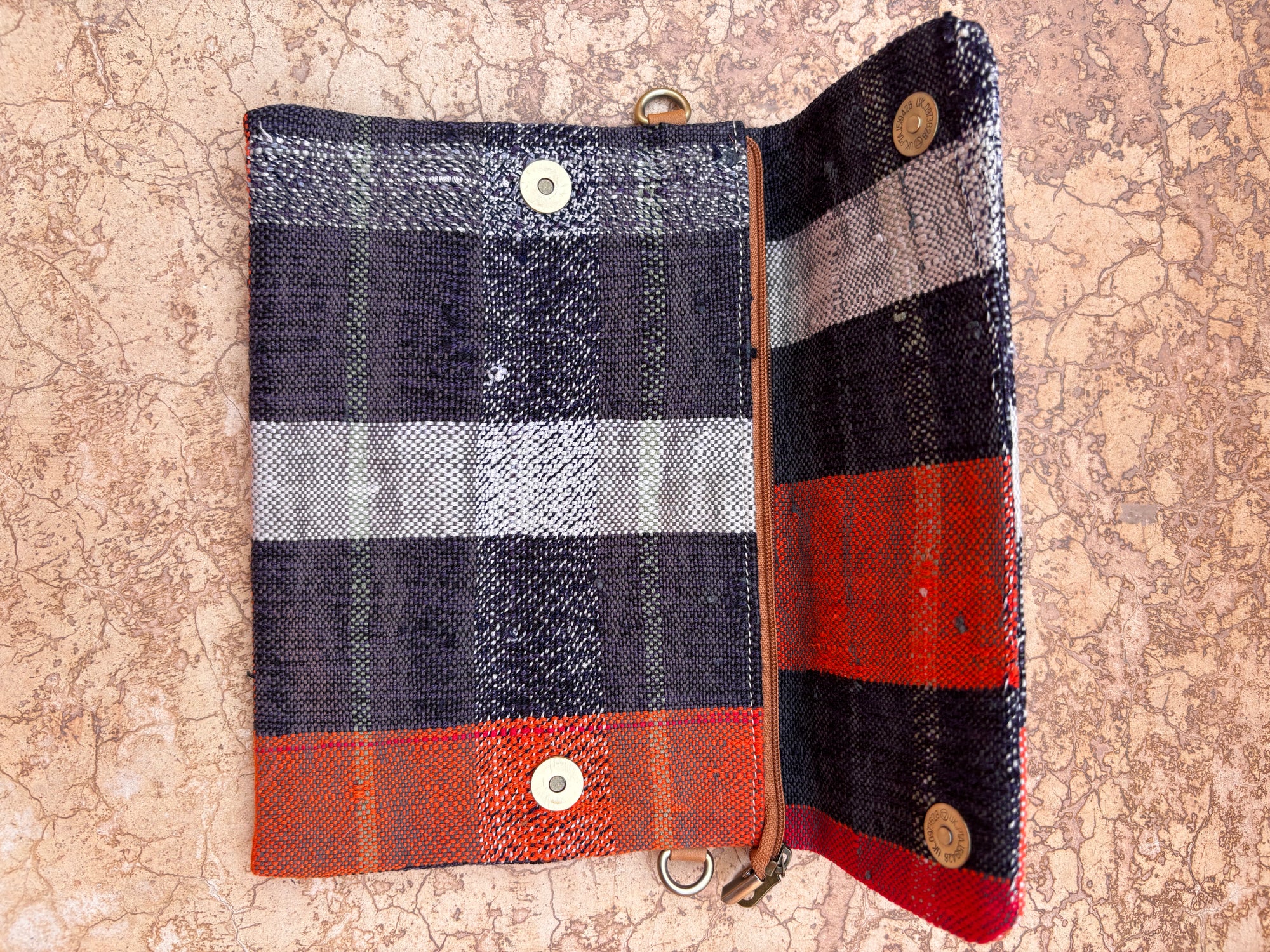 Die Vintage Clutch von LEY! ist eine handgefertigte Tasche aus gewebtem Stoff im Plaidmuster (Navy, Weiß, Grau, Rot, Orange) mit Druckknöpfen und Reißverschlussfach - abgebildet auf strukturiertem Stein.