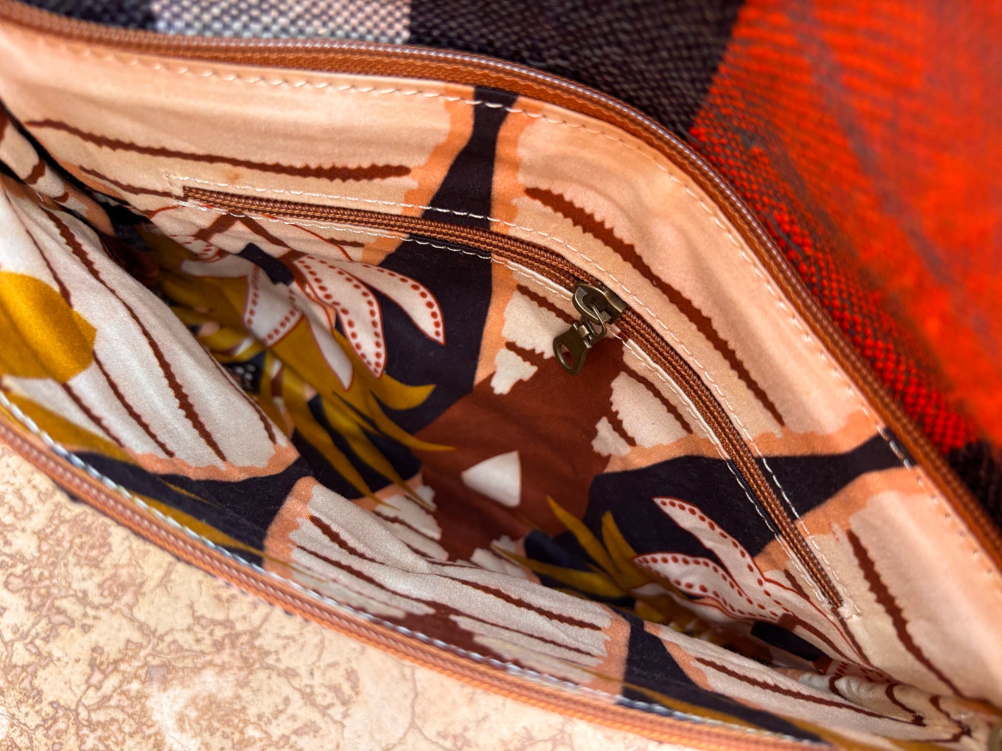 Nahaufnahme der LEY! Vintage Clutch mit sichtbaren Nähten, einem blumengemusterten Stofffutter und einer Reißverschlusstasche entlang des Lederbesatzes, dargestellt auf einer strukturierten beigen Oberfläche.