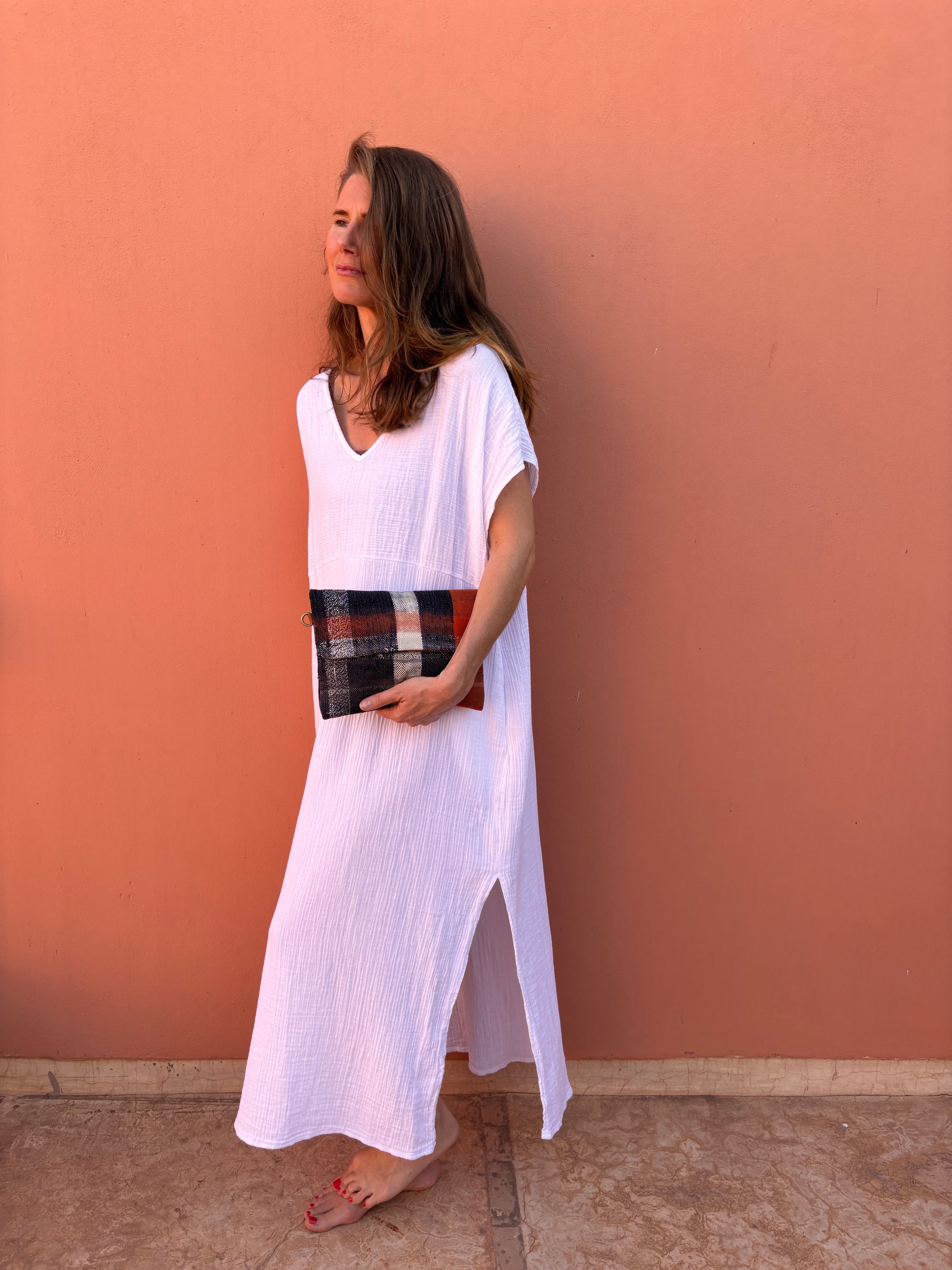 Eine Frau steht barfuß vor einer pfirsichfarbenen Wand und lächelt sanft, während sie die LEY! Vintage Clutch in der Hand hält und sie mit einem lockeren weißen, knöchellangen Kleid kombiniert.