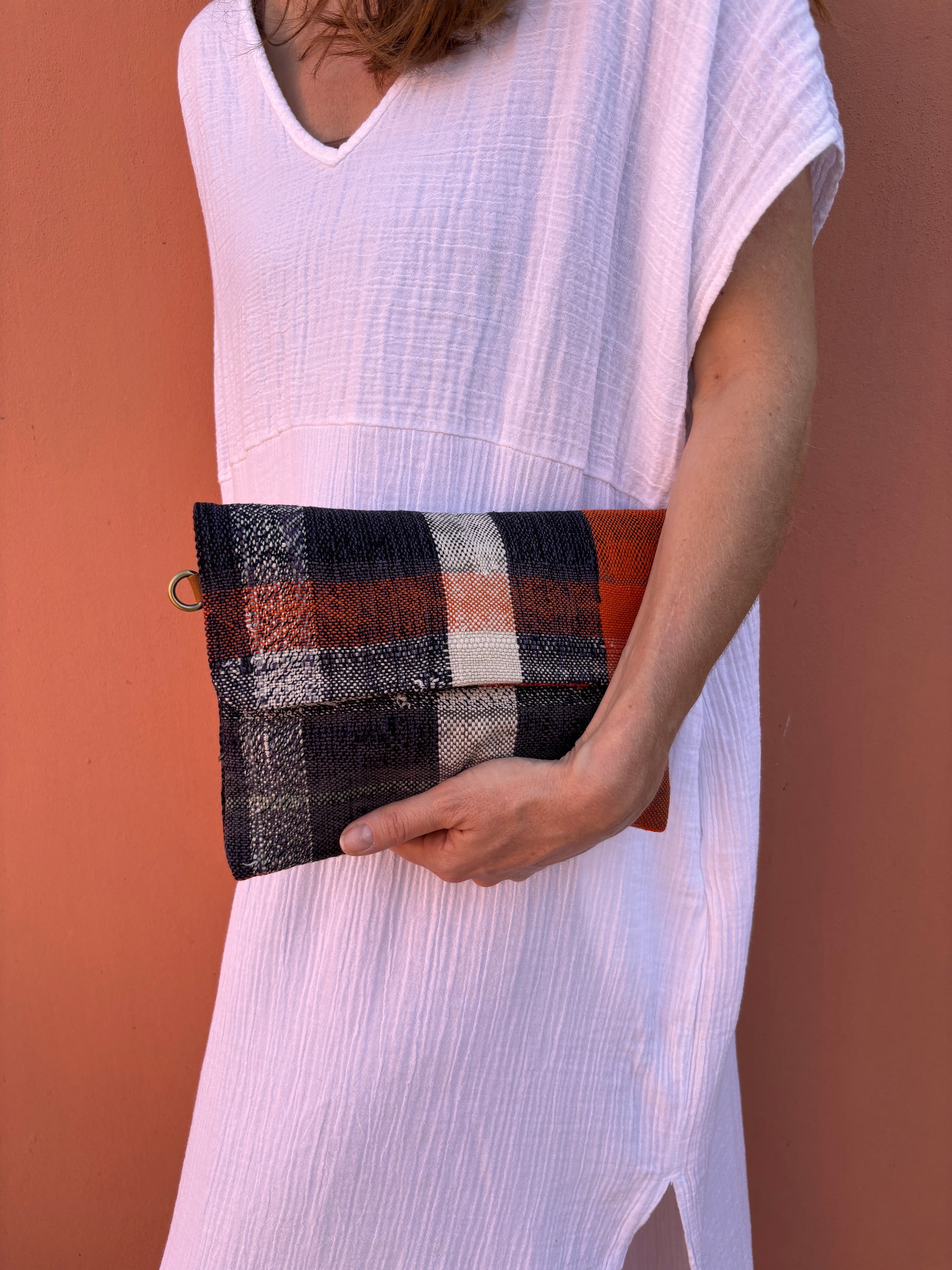 Eine Frau in einem weißen, strukturierten Kleid steht vor einer pfirsichfarbenen Wand und hält die LEY! Vintage Clutch - ein rechteckiges, kariertes Accessoire mit orangefarbenen, schwarzen und weißen Streifen - unter ihrem Arm.