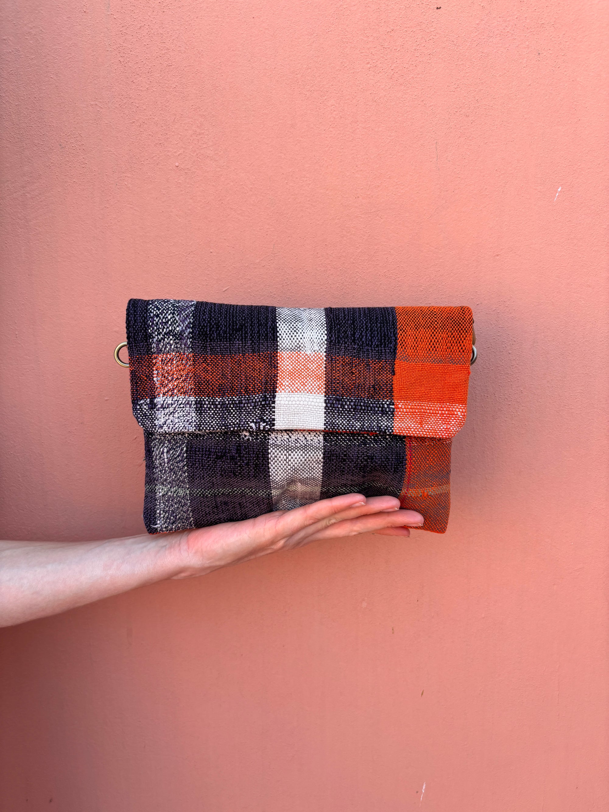 Eine Hand hält die LEY! Vintage Clutch und präsentiert ihr auffälliges Karomuster in Orange, Weiß, Schwarz und Marineblau vor einer glatten pfirsichfarbenen Wand.