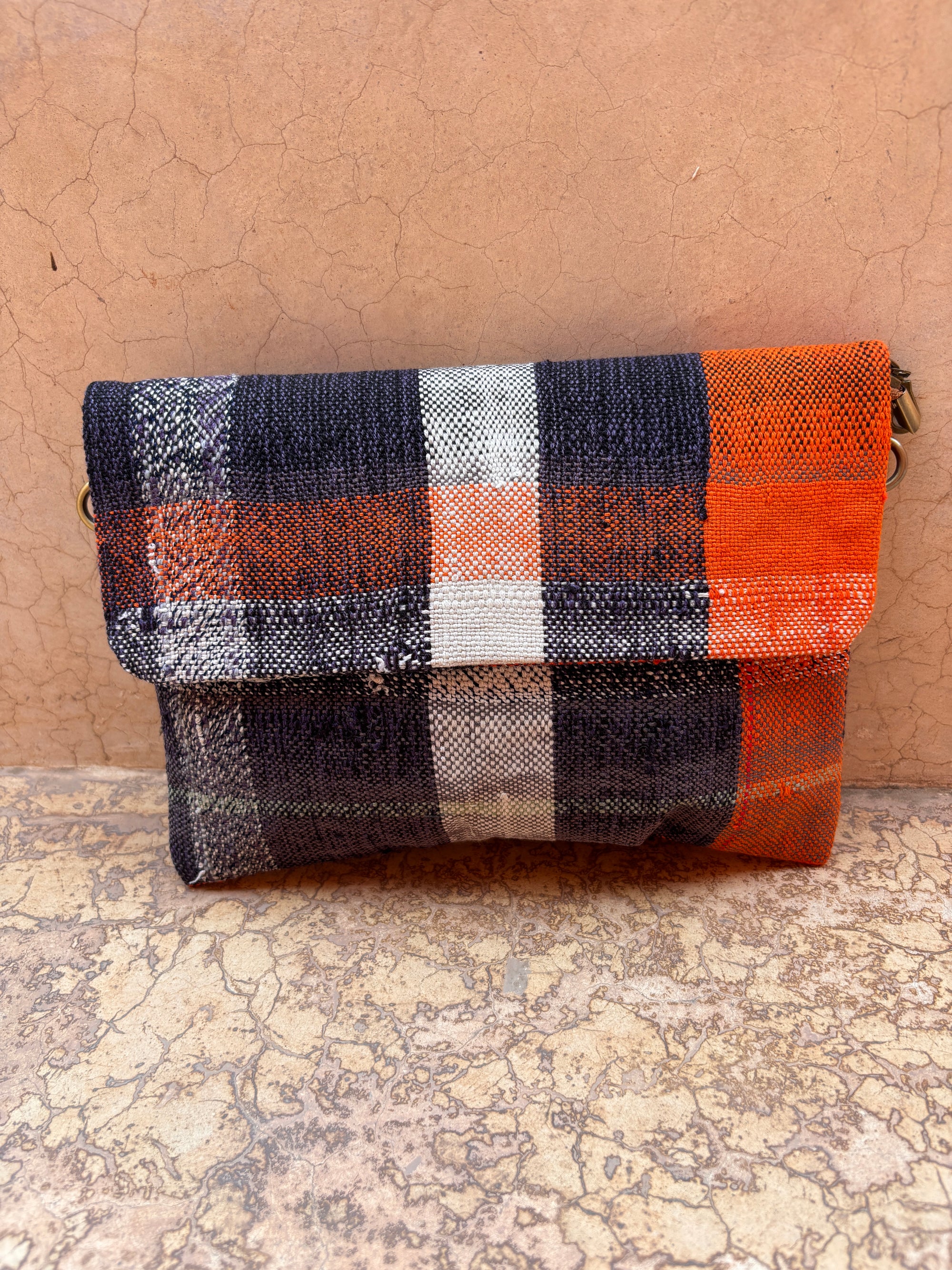 Die LEY! Vintage Clutch, eine kleine rechteckige Tasche mit Überschlag und auffälligem Karomuster in Marineblau, Orange, Weiß und Schwarz, wird vor einer beigefarbenen Wand und einem beigen Boden präsentiert.