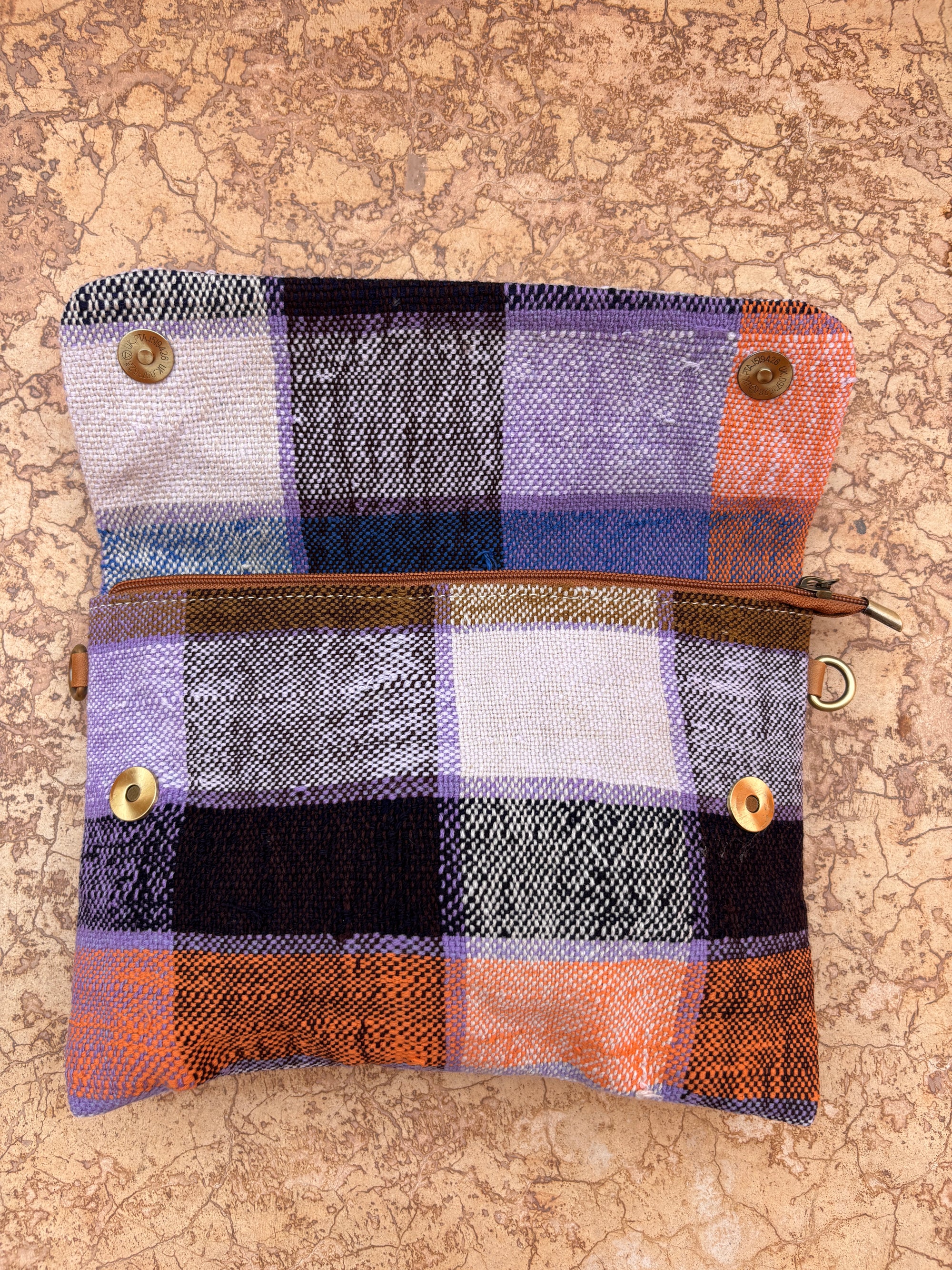 Die LEY! Vintage Clutch ist eine handgefertigte Stofftasche mit braunem Reißverschluss und Druckknöpfen, die ein lila, schwarz, orange und weiß kariertes Design zeigt - ideal als stilvolles Accessoire auf jeder strukturierten Oberfläche.