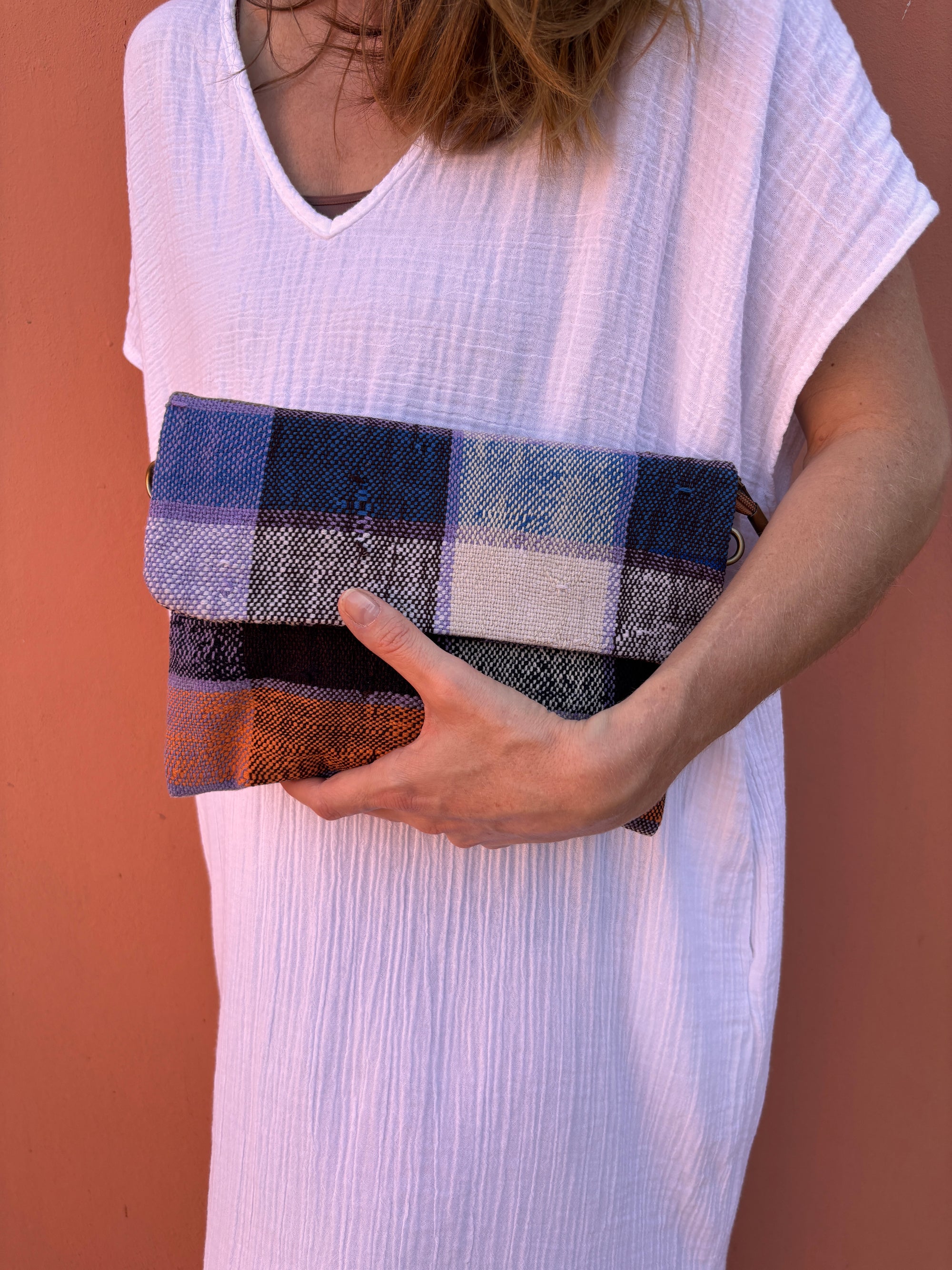 Eine Person in einem strukturierten weißen Kleid hält die LEY! Vintage Clutch, eine handgefertigte, upgecycelte Tasche mit blauem, schwarzem, weißem und orangefarbenem Karomuster, vor einer pfirsichfarbenen Wand.